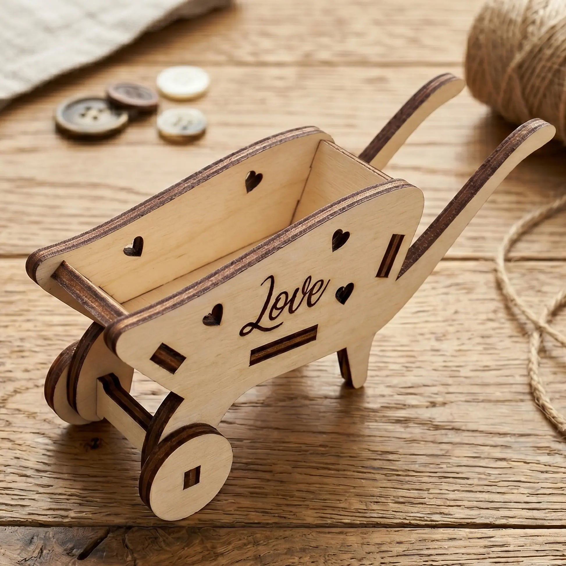 Miniature laser cut plywood wheelbarrow with heart cutouts and "Love" engraving on a rustic wooden desk with sewing buttons