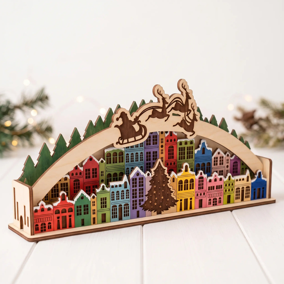 Front angle view of painted laser cut plywood Christmas town arch diorama on white table with festive greenery and lights.