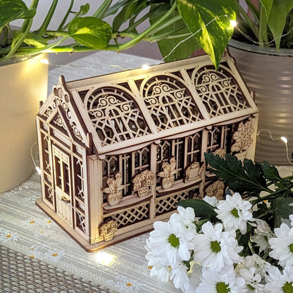 Ornate miniature greenhouse crafted from laser cut plywood, illuminated by string lights next to white fresh flowers.