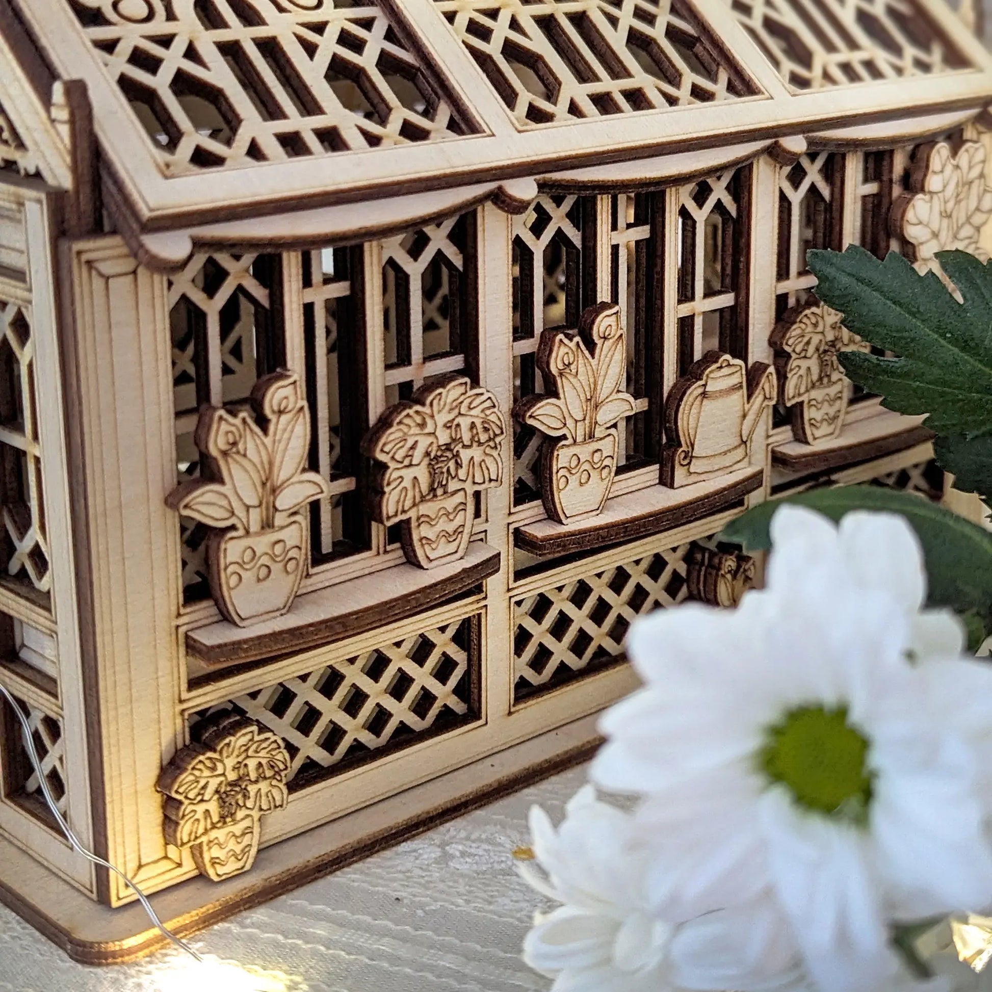 Close-up of a greenhouse made of laser cut plywood, featuring window shelves with tiny potted plants and a watering can.