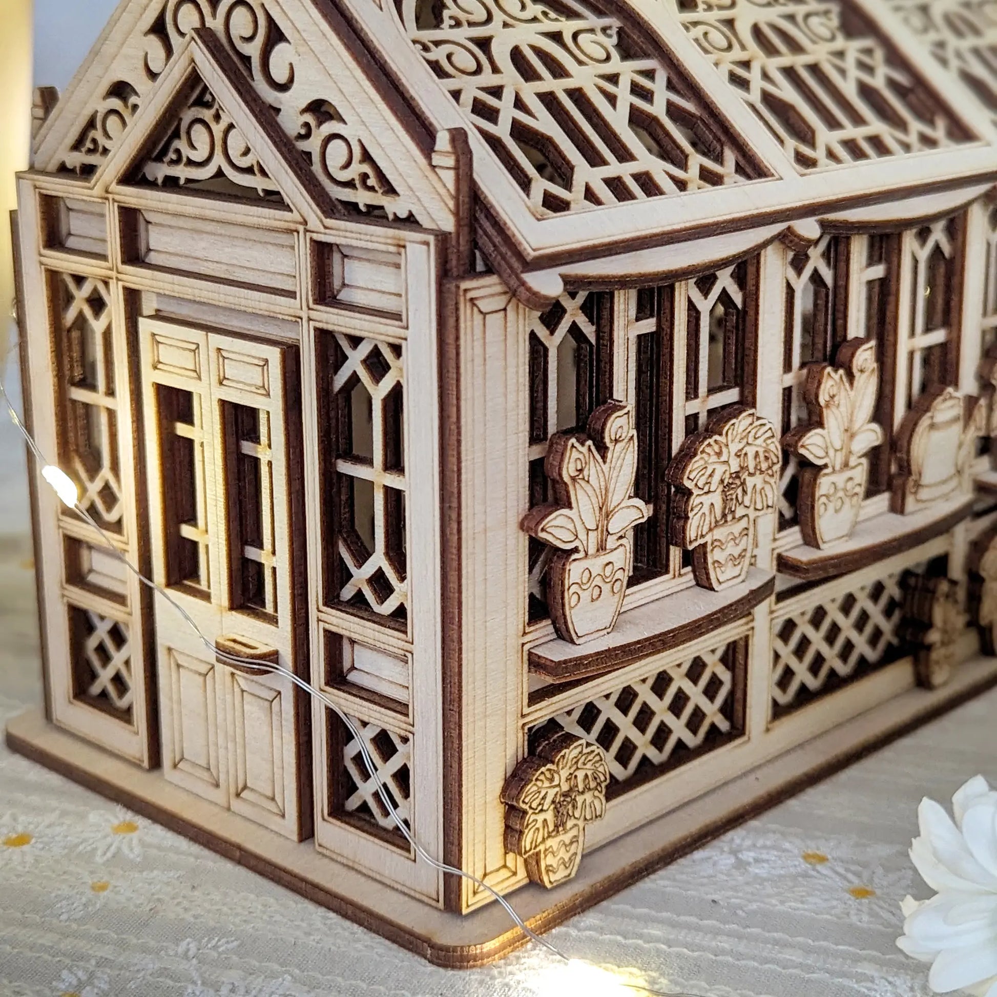 Side view of laser cut plywood miniature house with intricate carved doors, windows, and potted plant silhouettes.