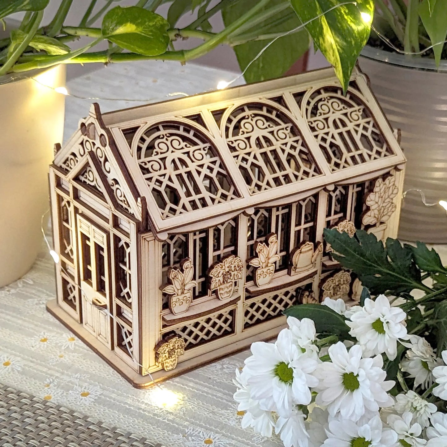 Top-angle view of a decorative laser cut plywood greenhouse with ornate arched roof panels next to white blossoms.