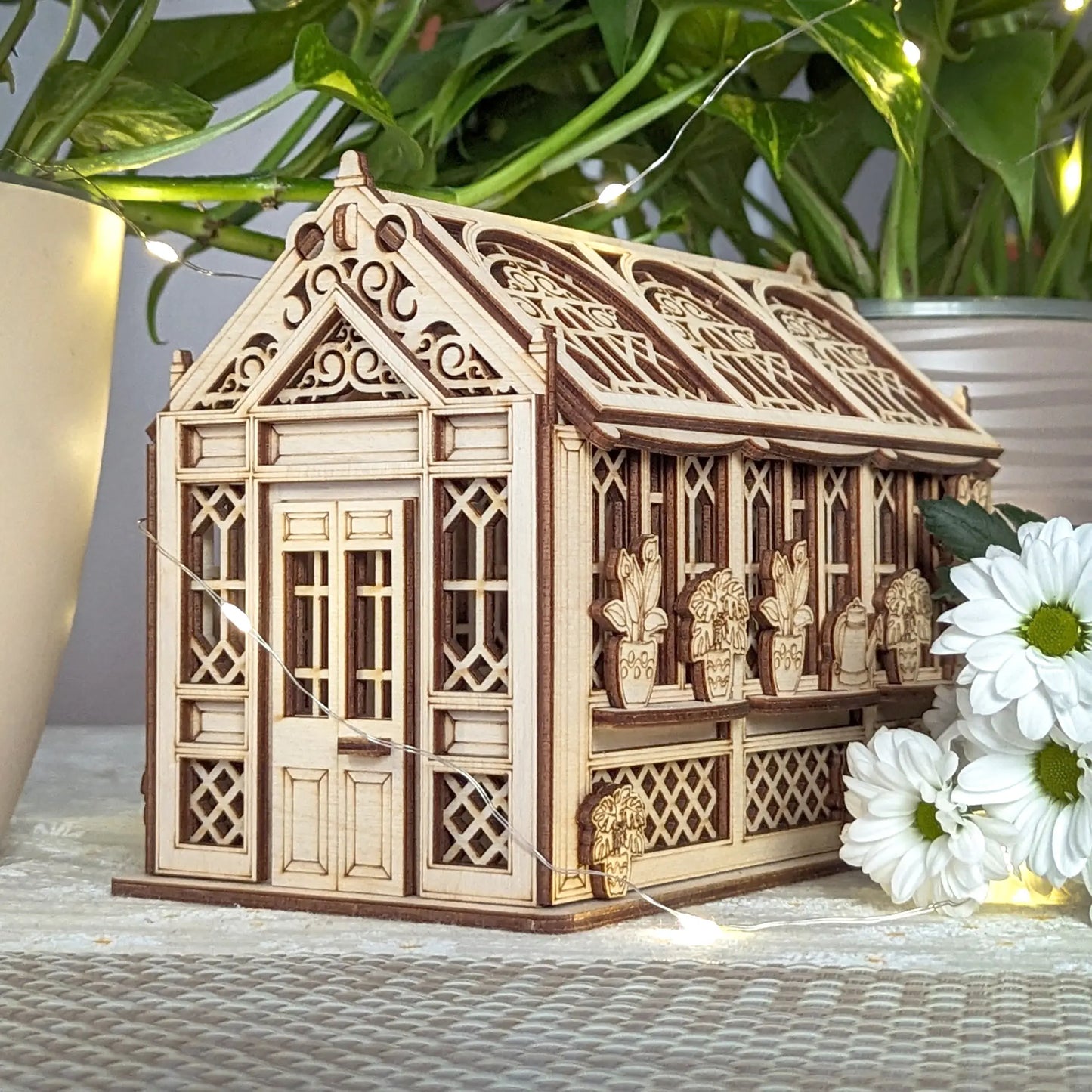 Miniature laser cut plywood greenhouse with gothic carved roof, surrounded by fairy lights and white daisy flowers.