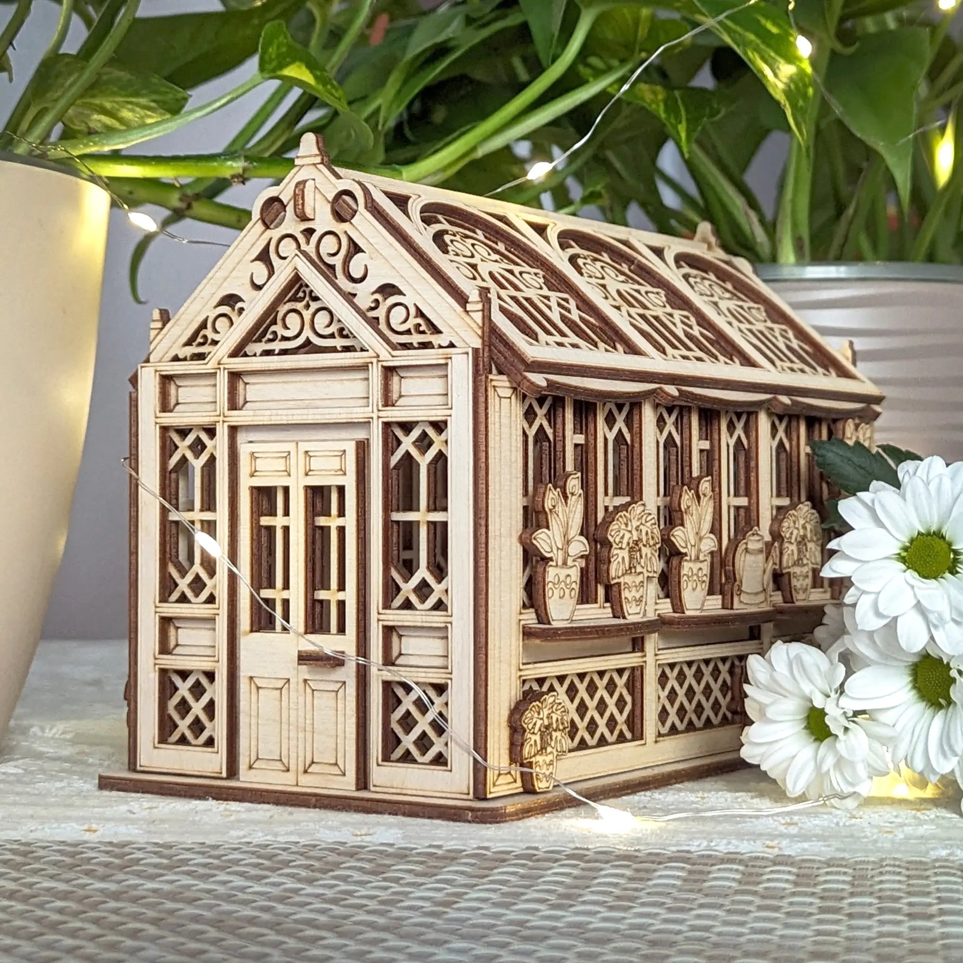 Miniature laser cut plywood greenhouse with gothic carved roof, surrounded by fairy lights and white daisy flowers.