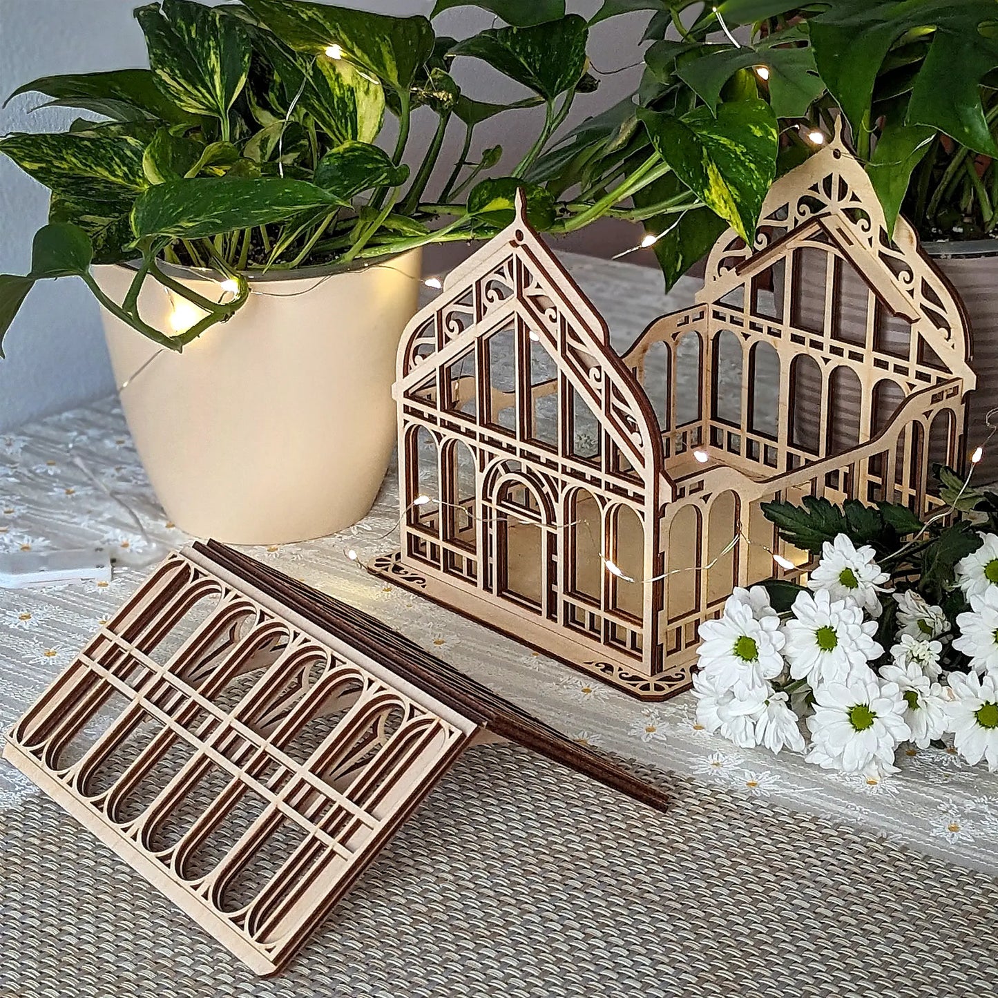 Miniature laser cut plywood greenhouse with its roof removed, sitting next to potted plants and white daisies.