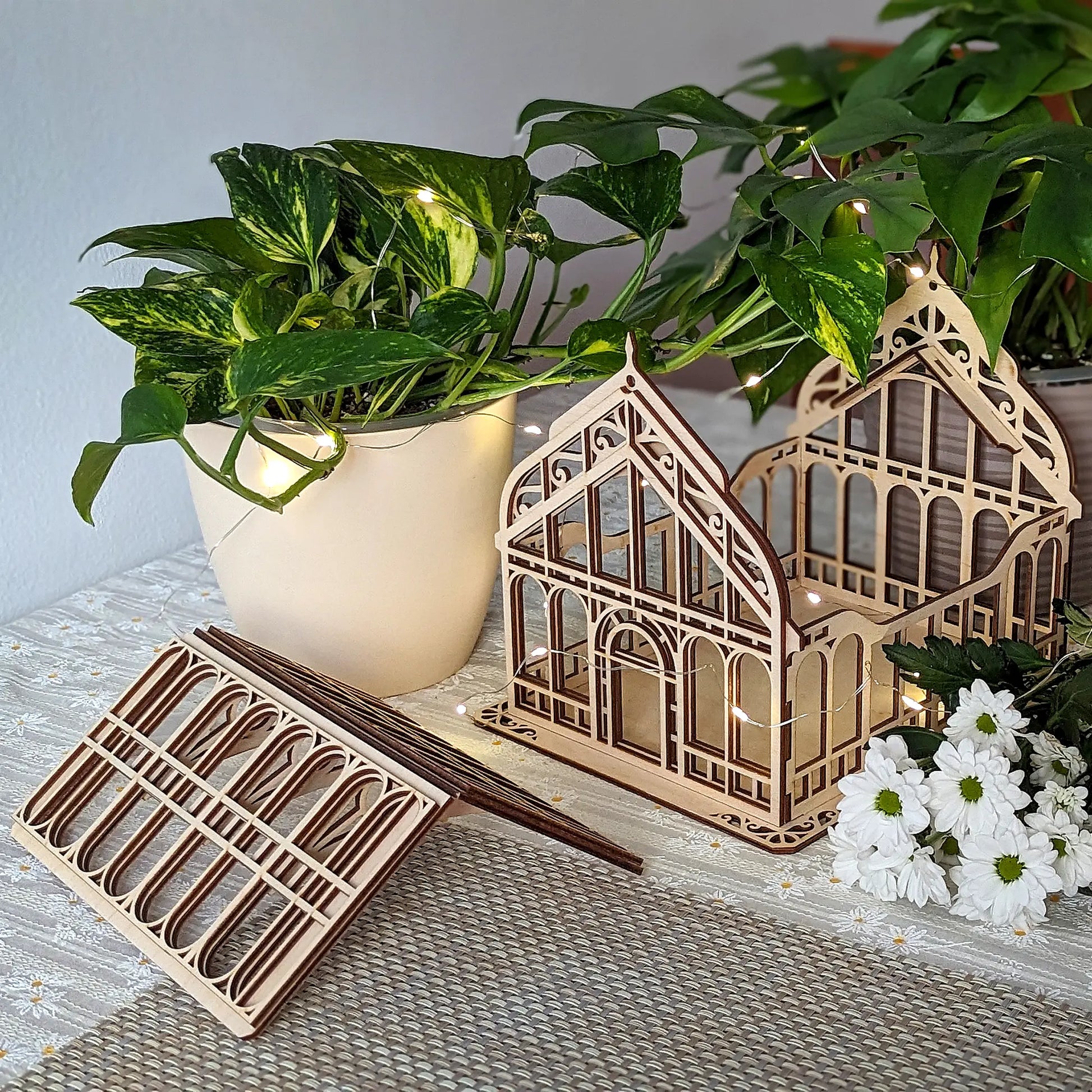 Assembled laser cut plywood dollhouse greenhouse with a detached roof on a tablecloth near a flower bouquet.