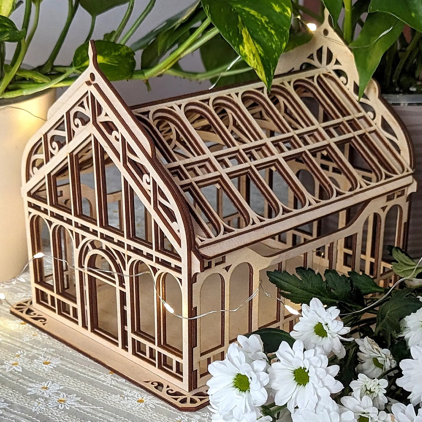 Elegant laser cut plywood miniature greenhouse box with Gothic arches, lit by warm lights next to white daisies.