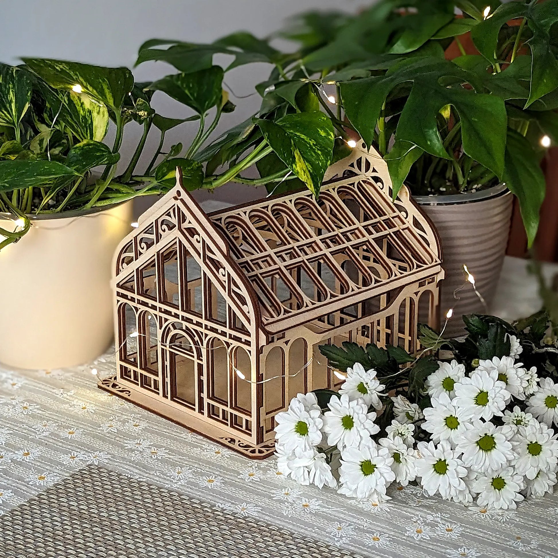 Miniature Victorian greenhouse of laser cut plywood on a table among green house plants and white flowers.