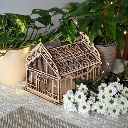 Miniature Victorian greenhouse of laser cut plywood on a table among green house plants and white flowers.