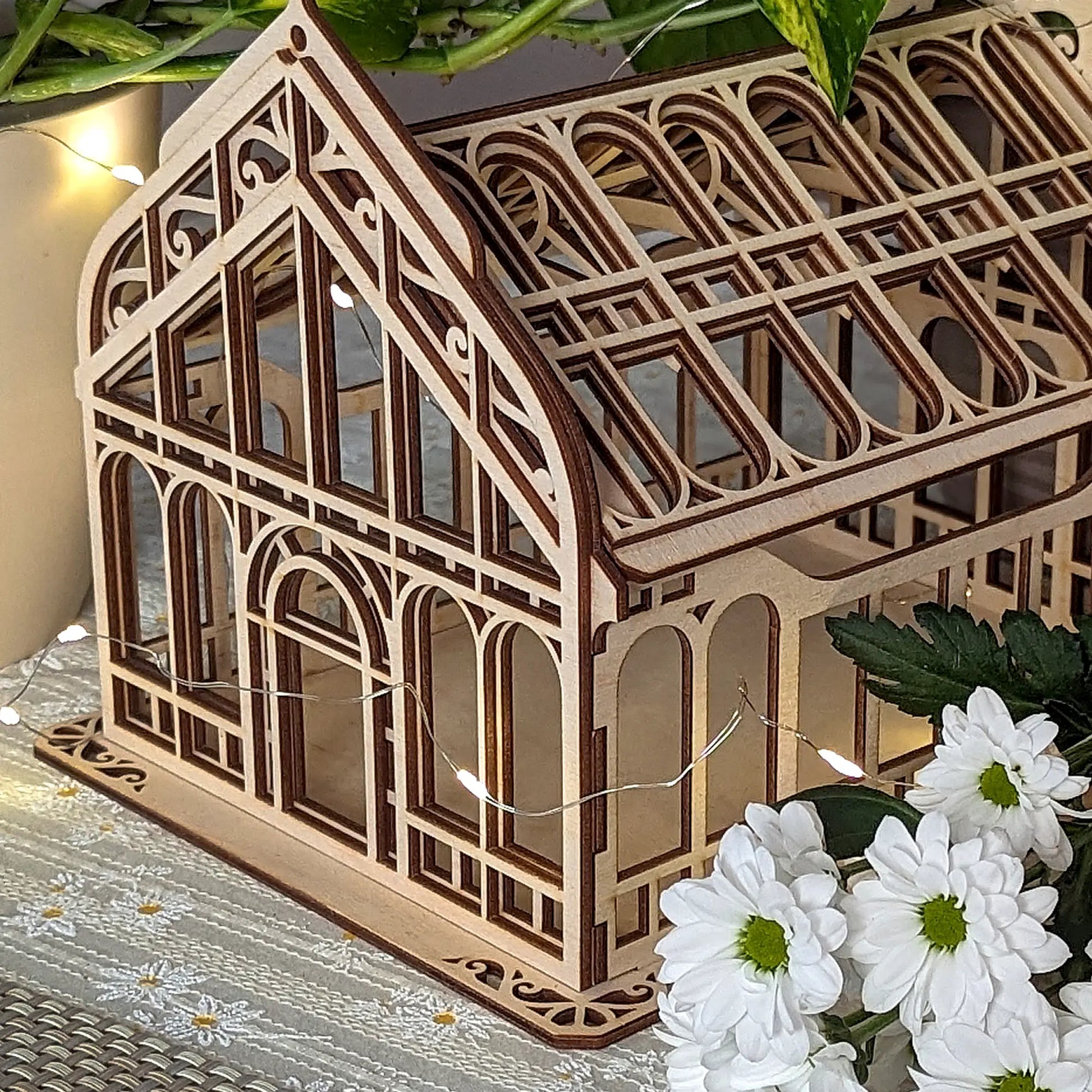 Close-up of a laser cut plywood miniature greenhouse decorated with a fairy light string and white flowers.