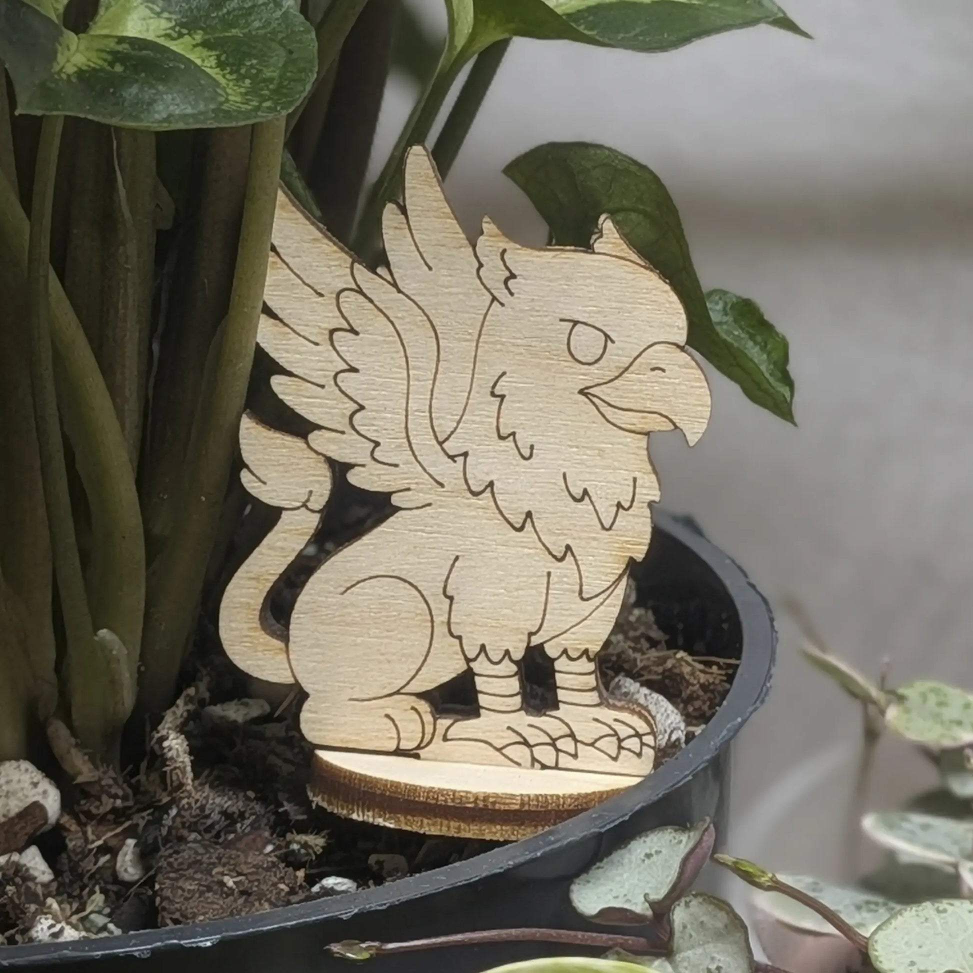 Laser cut plywood griffin figurine sits in a plant pot. Features eagle wings, lion body, and fine engraved feather details