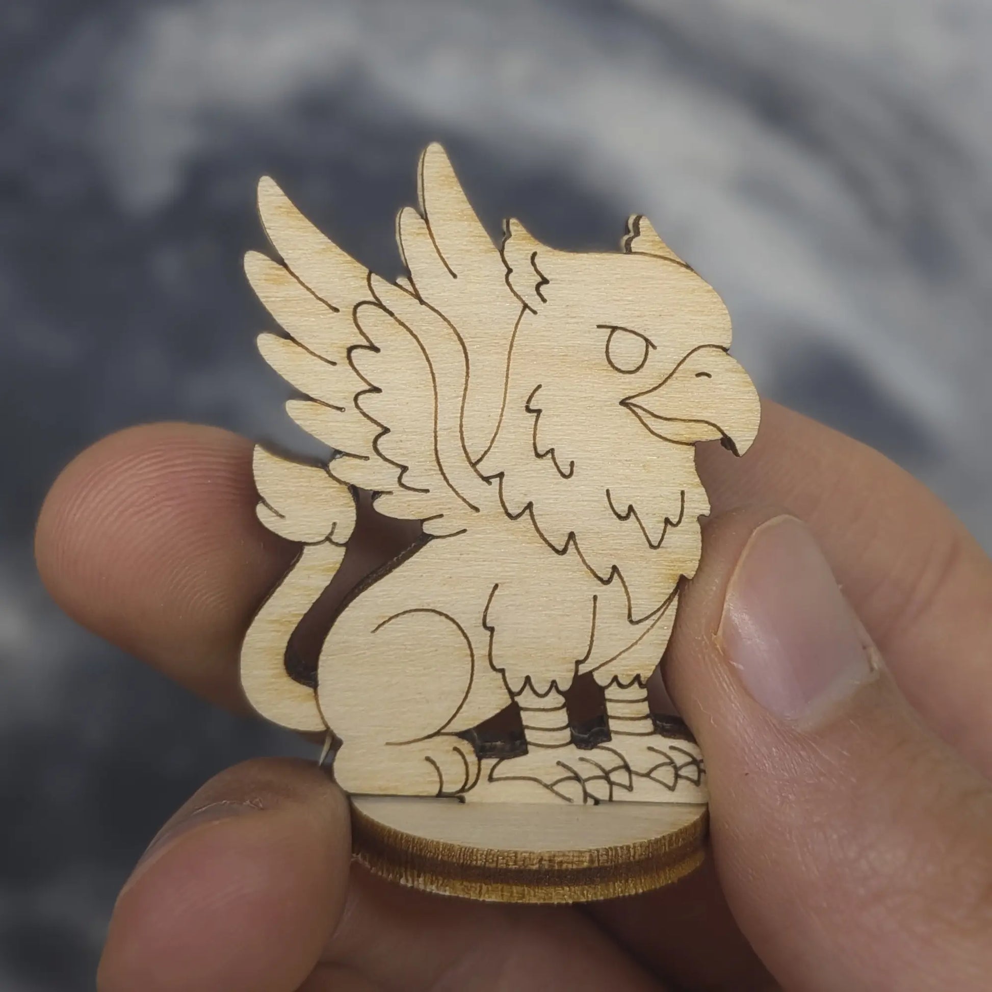 Hand holds a laser cut plywood griffin miniature. Showing eagle beak, sharp talons, and layered wing engravings for scale