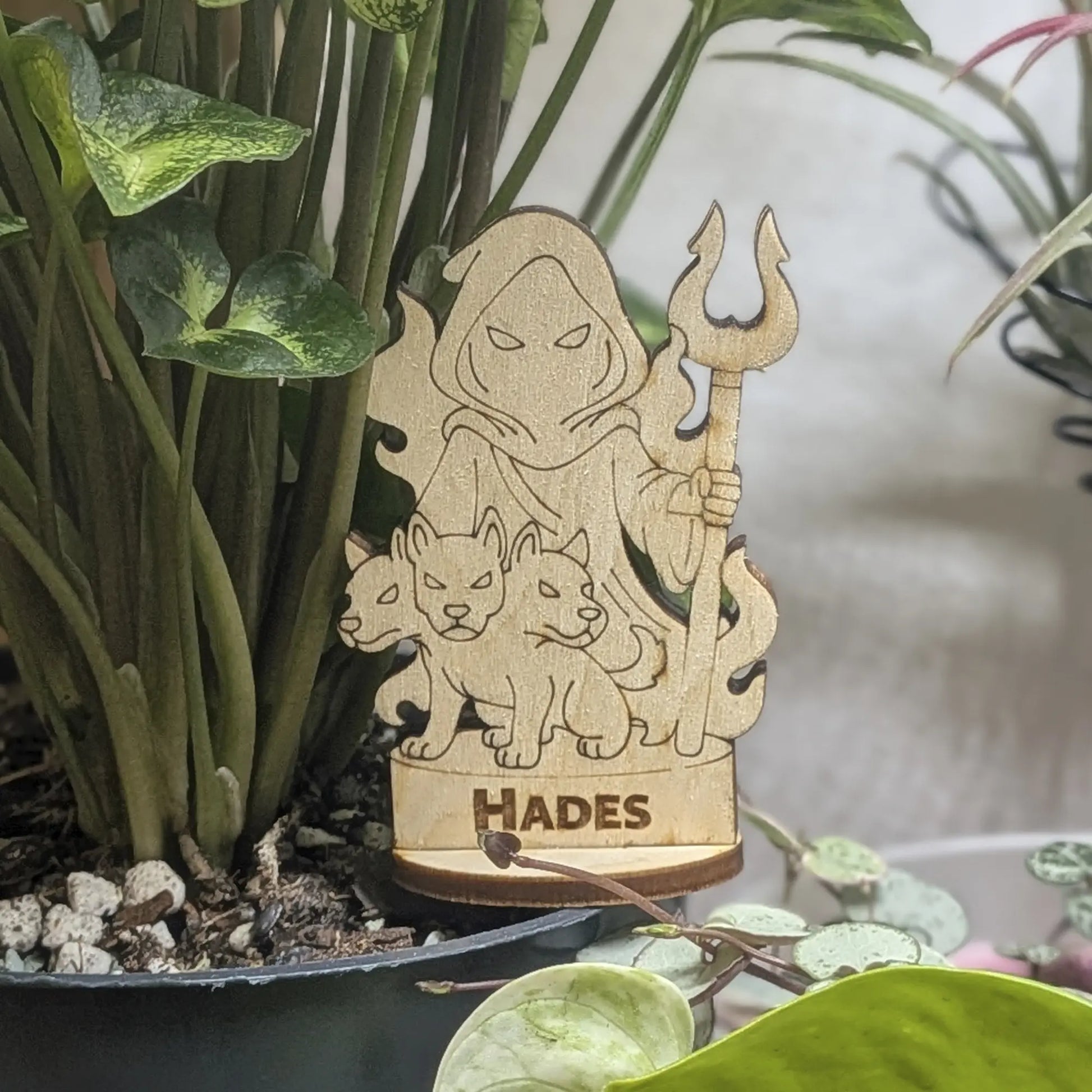 Laser cut plywood Hades figurine sits in a plant pot. Features a crown, bident staff, and detailed robes on wood base