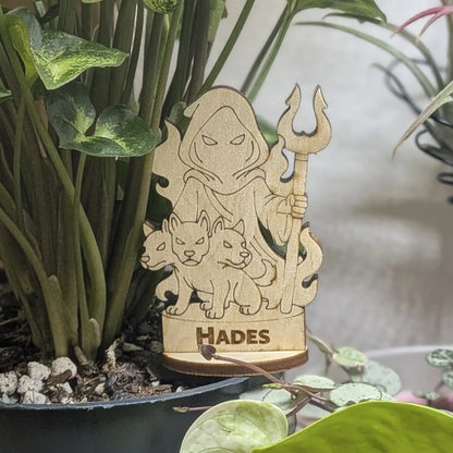 Laser cut plywood Hades figurine sits in a plant pot. Features a crown, bident staff, and detailed robes on wood base