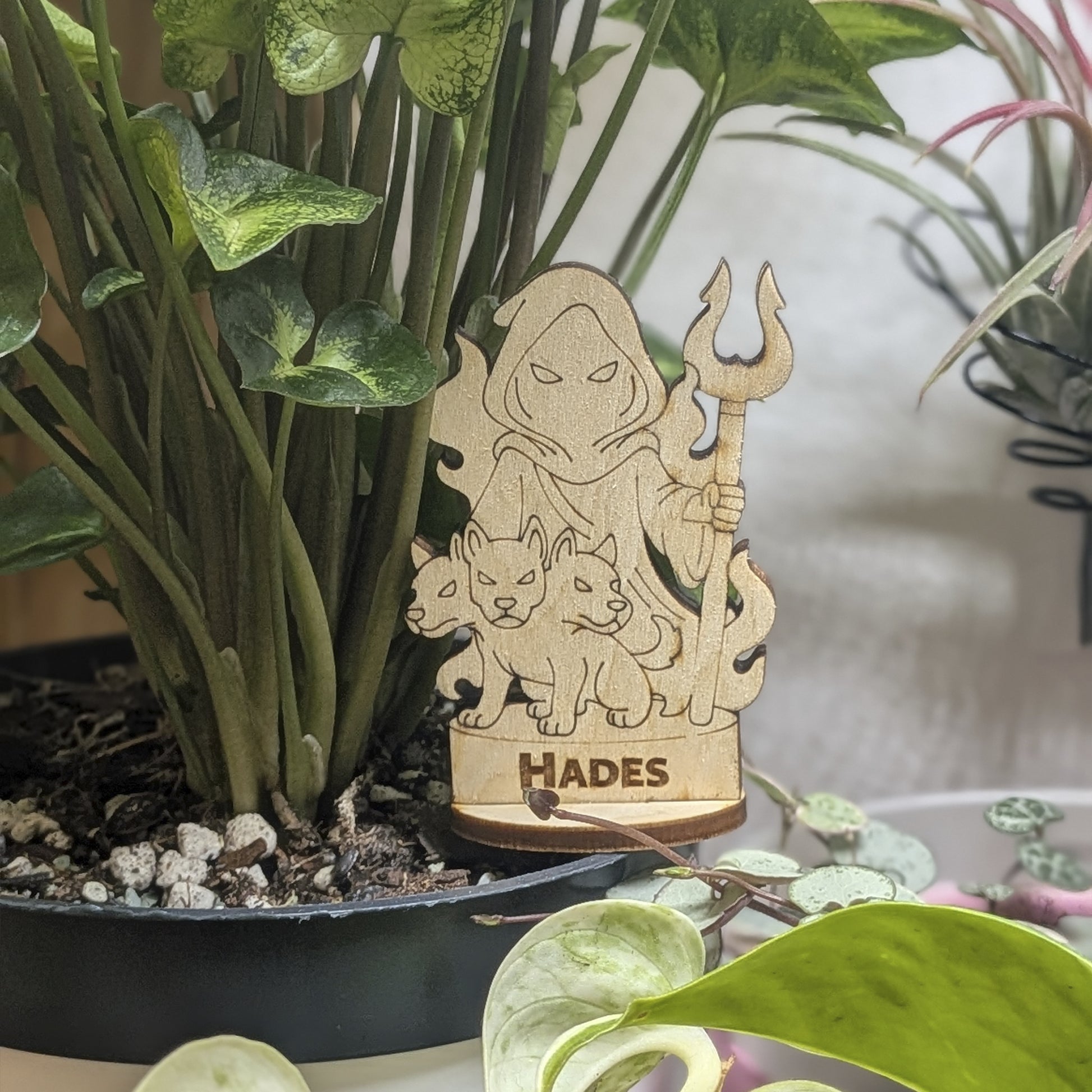 Laser cut plywood Hades figurine sits in a plant pot. Features a crown, bident staff, and detailed robes on wood base