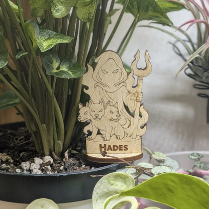 Laser cut plywood Hades figurine sits in a plant pot. Features a crown, bident staff, and detailed robes on wood base