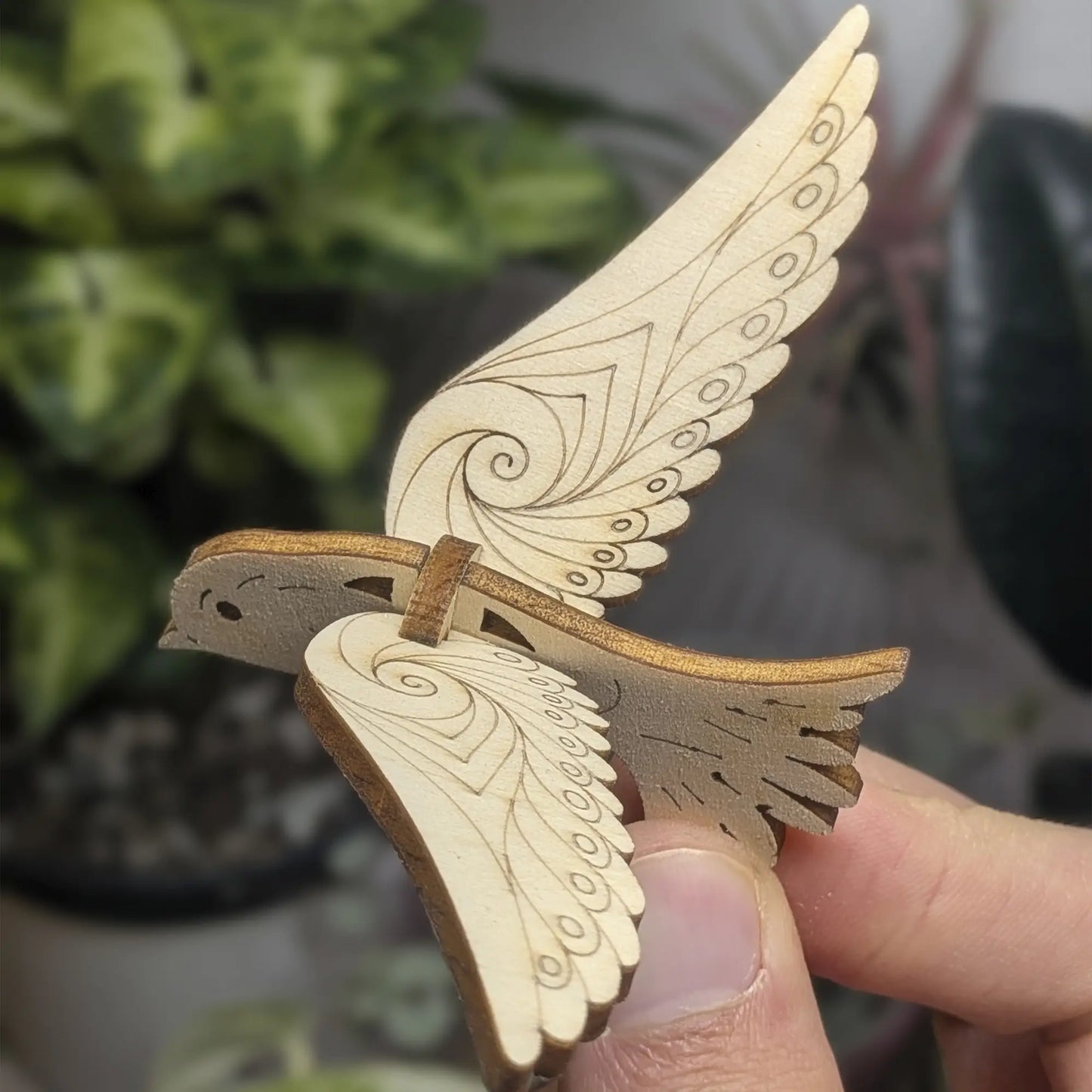 Hand holds a 3D bird ornament of laser cut plywood with wings raised. Detailed swirl engravings and dark burnt edges
