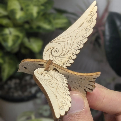 Hand holds a 3D bird ornament of laser cut plywood with wings raised. Detailed swirl engravings and dark burnt edges