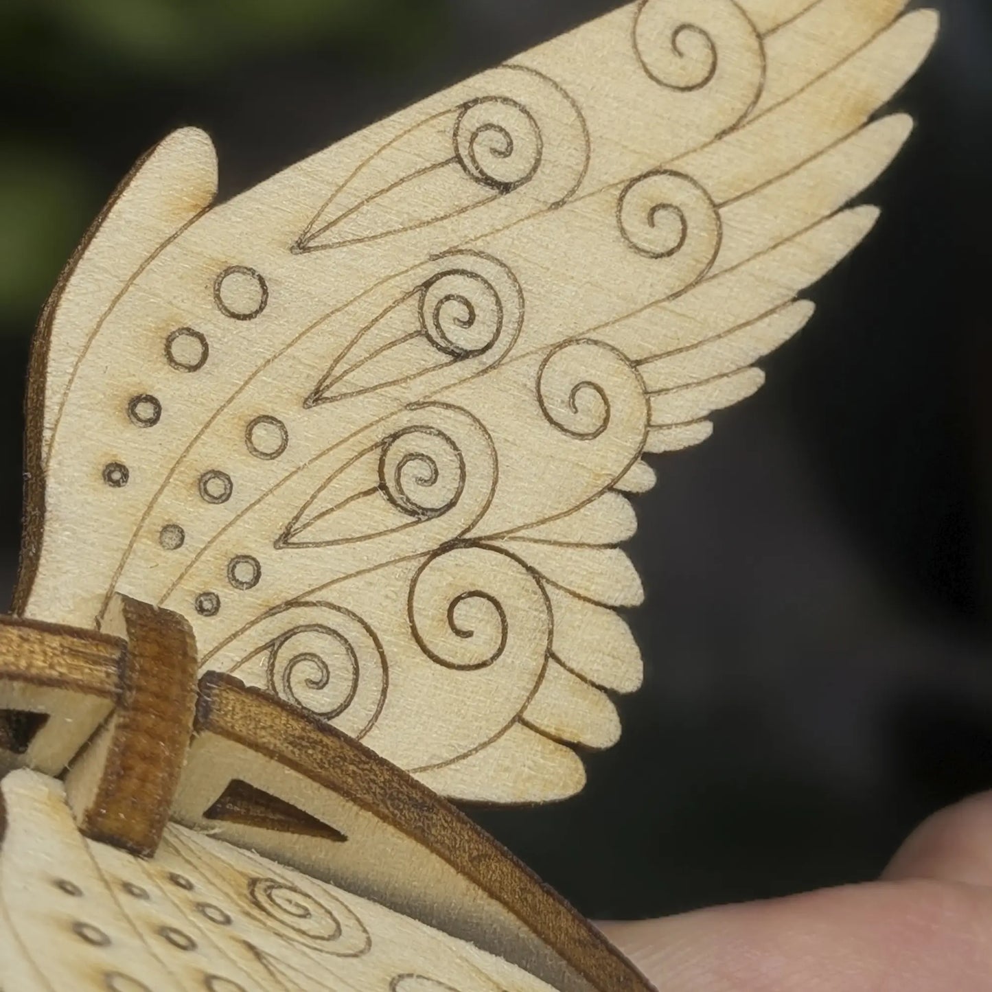Macro of a laser cut plywood bird wing showing dark burnt edges and intricate engraved patterns of swirls and circles.