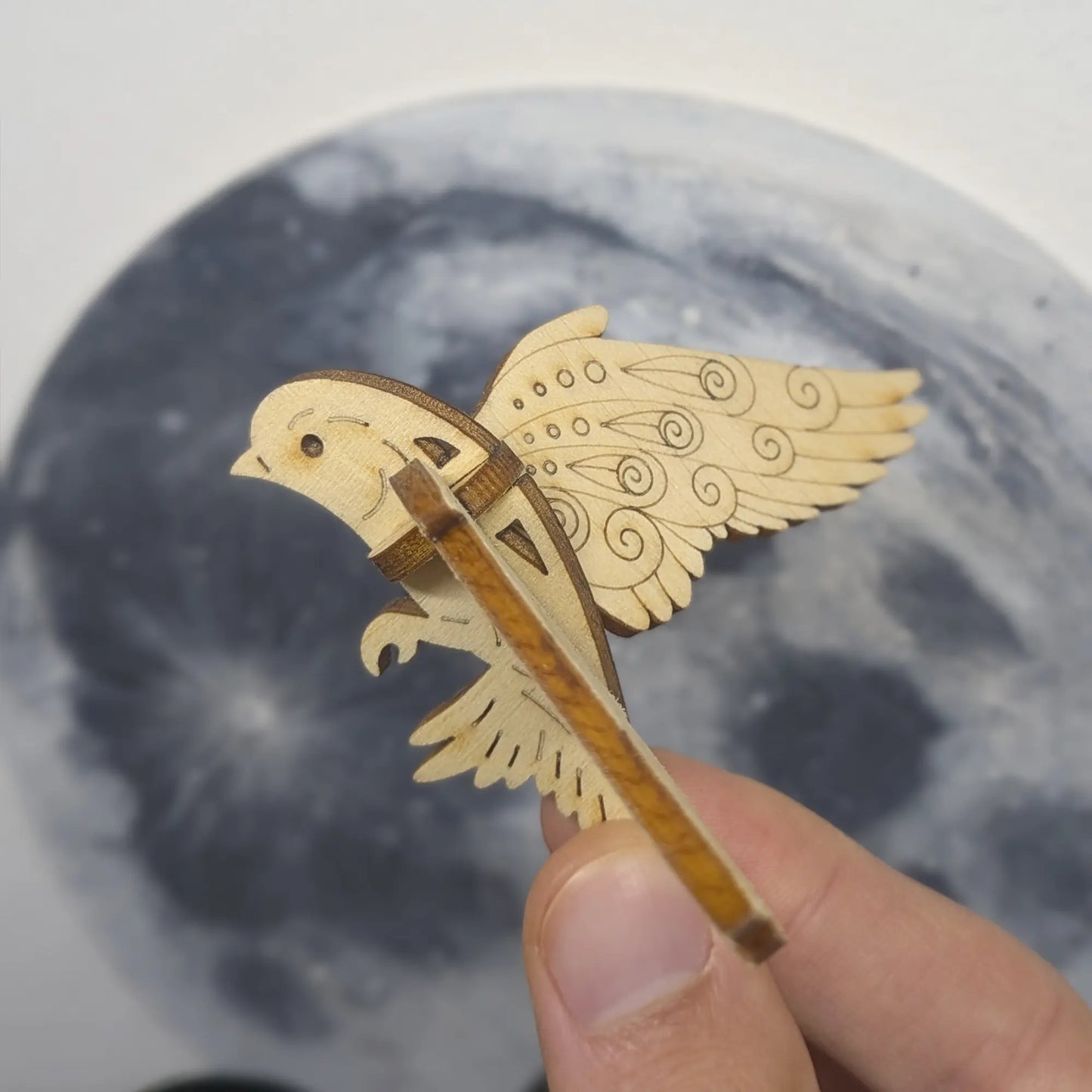 Hand holds a laser cut plywood bird ornament in flight. Side profile shows detailed wing etchings and a moon background