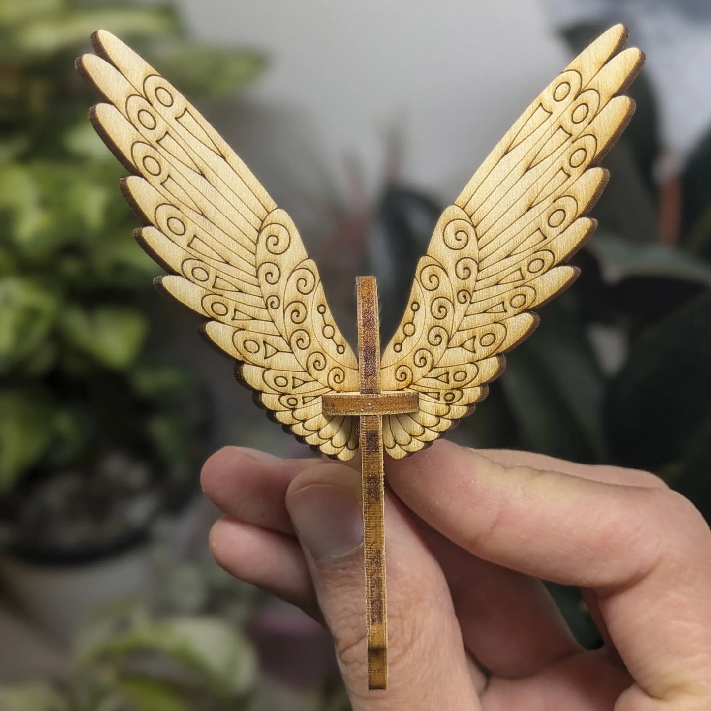 Hand holds a laser cut plywood bird with spiral wing patterns against green leaves.