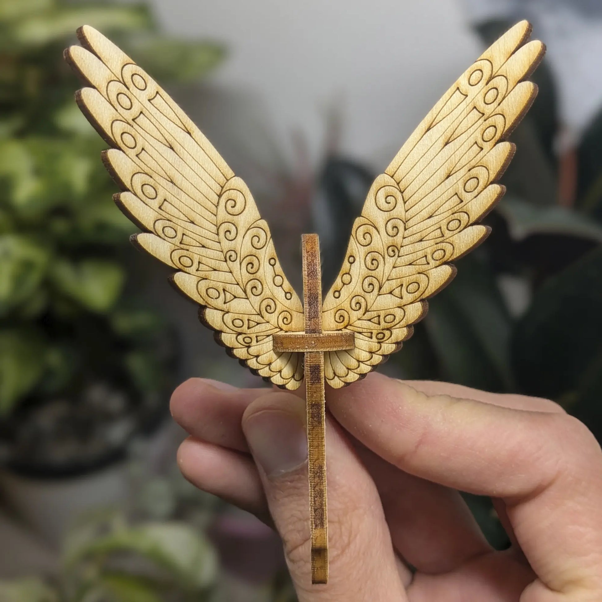 Hand holds a laser cut plywood bird with spiral wing patterns against green leaves.