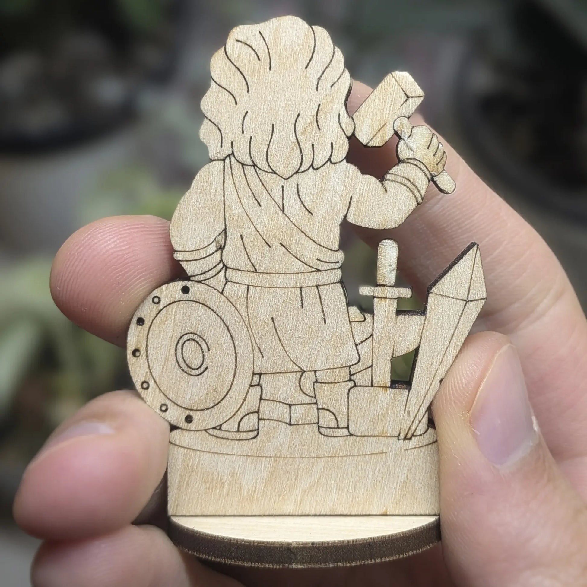 Back view of laser cut plywood Hephaestus held in hand. Shows dark burnt wood edges and detailed engraving on his cloak