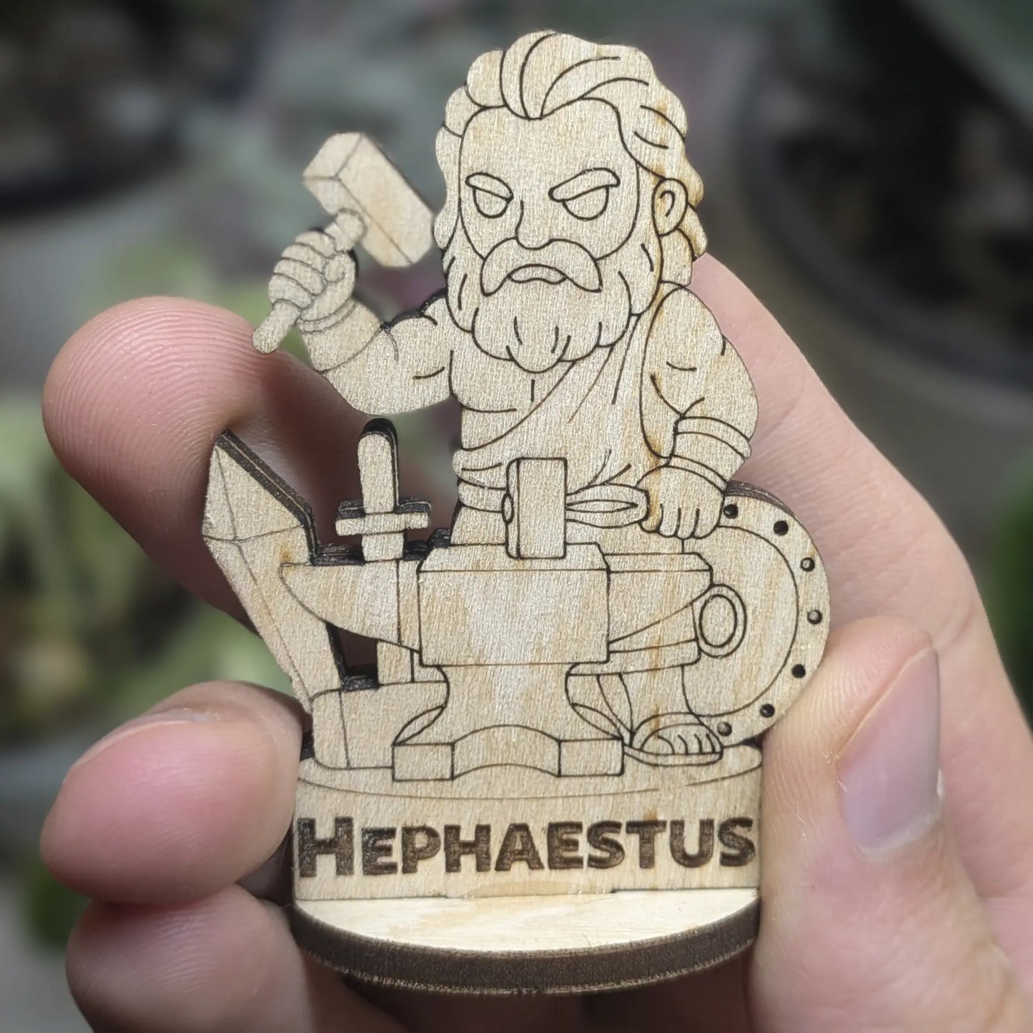 Hand holds a laser cut plywood Hephaestus miniature. Front view shows a bearded smith with a hammer and pliers for scale
