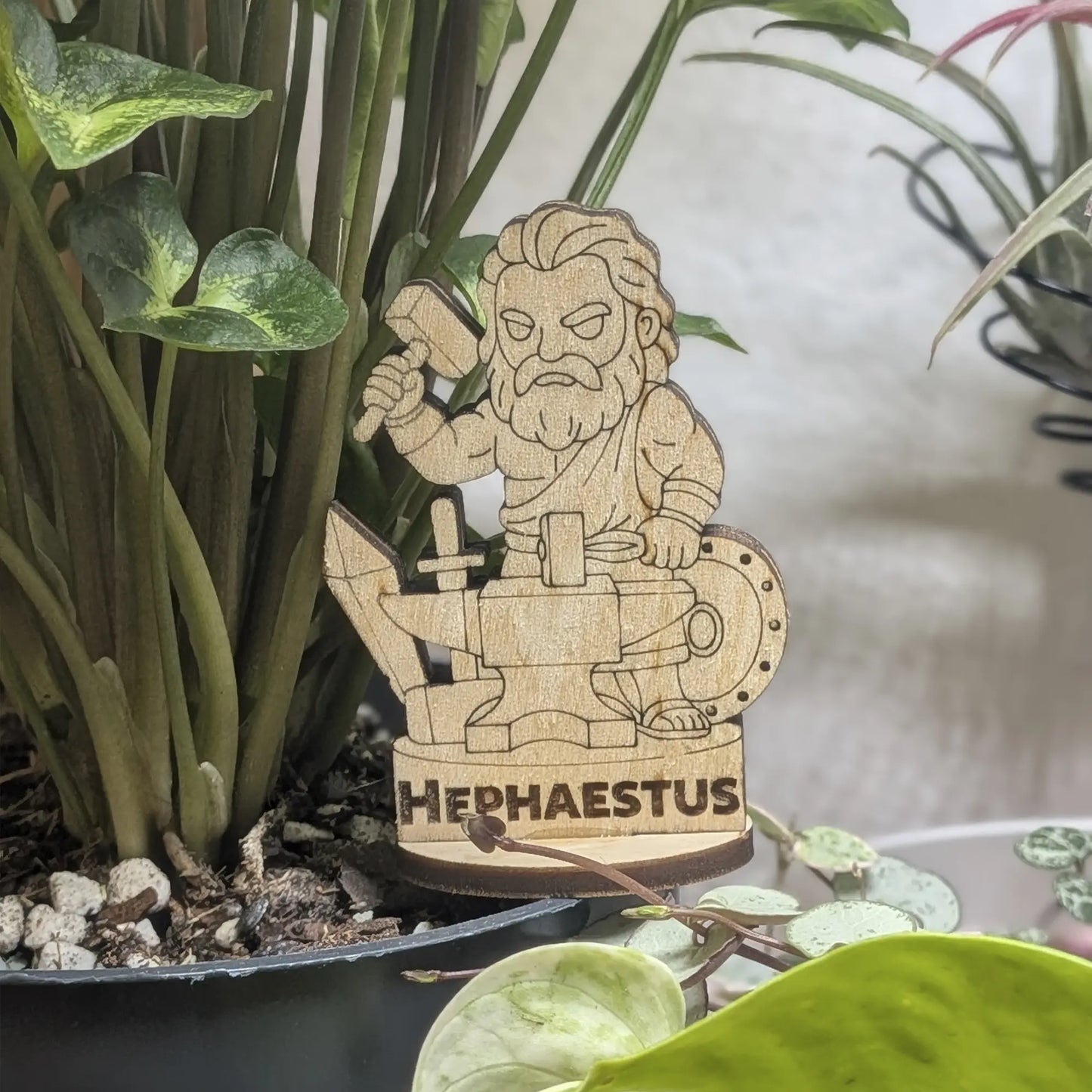 A laser cut plywood Hephaestus figurine in a plant pot. Blacksmith god with a hammer and tongs, featuring fine engraving