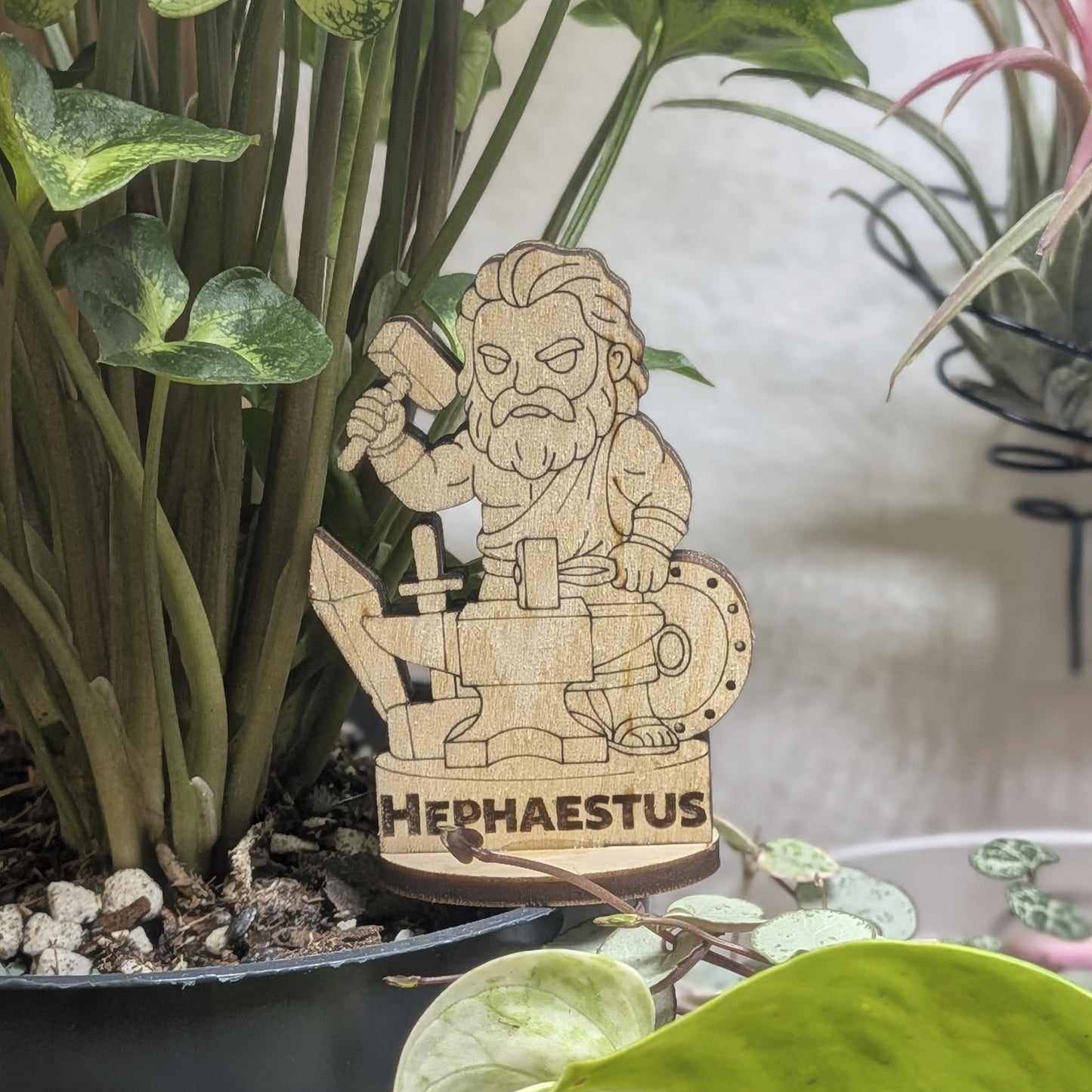 A laser cut plywood Hephaestus figurine in a plant pot. Blacksmith god with a hammer and tongs, featuring fine engraving