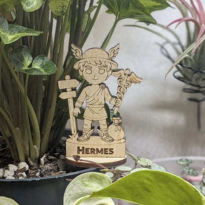 Laser cut plywood Hermes figurine sits in a plant pot. Features a caduceus staff, winged helmet, and shoes on a wood base