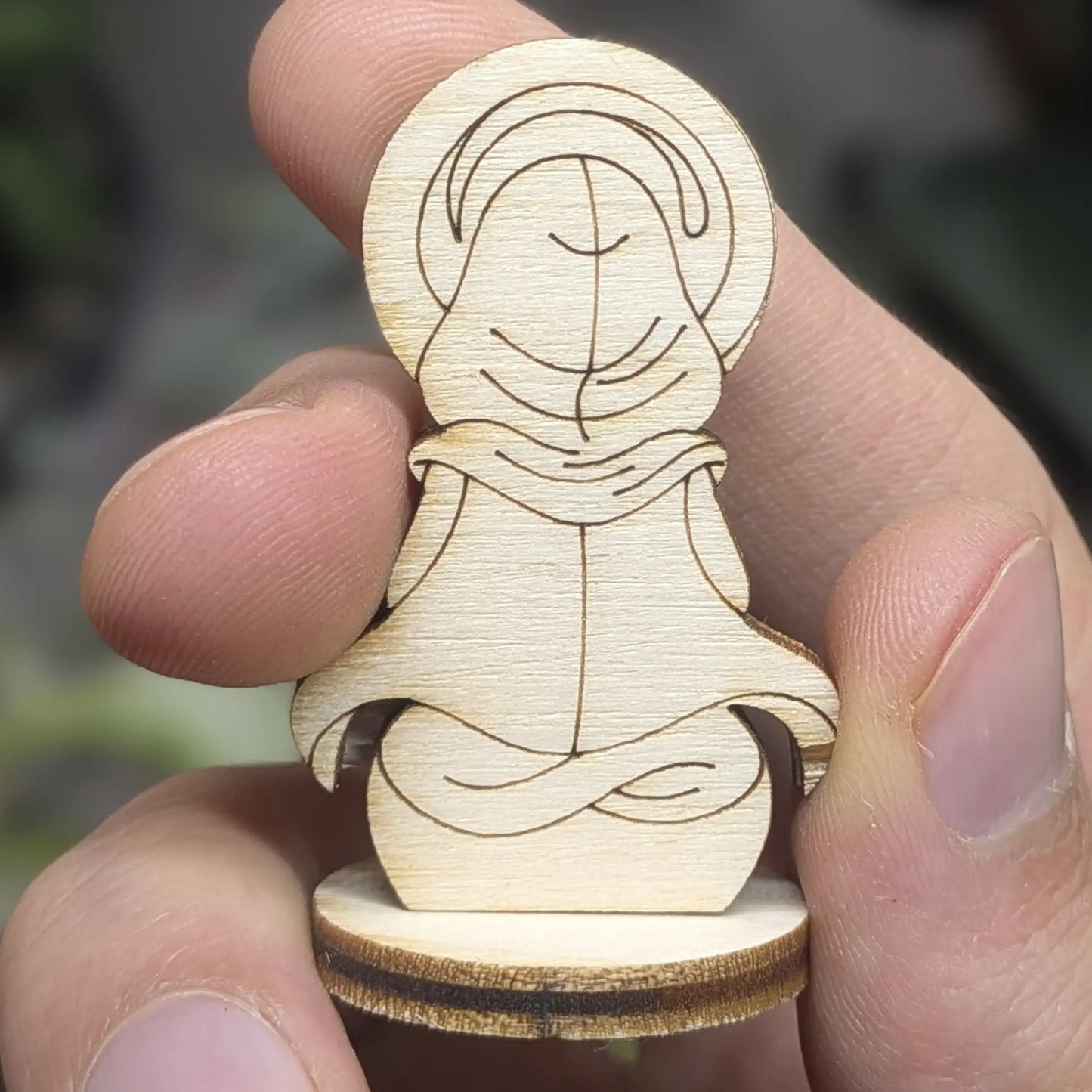 Back view of a holy monk figurine of laser cut plywood held in hand, showing a hooded robe and dark burnt wood grain edges