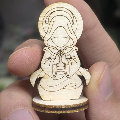 Hand holds a holy monk figurine of laser cut plywood, showing praying hands, a hooded cloak, and detailed halo engravings