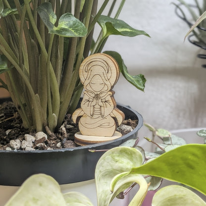 A holy monk figurine of laser cut plywood sits in a plant pot, featuring a meditating pose with hands together and a halo ring