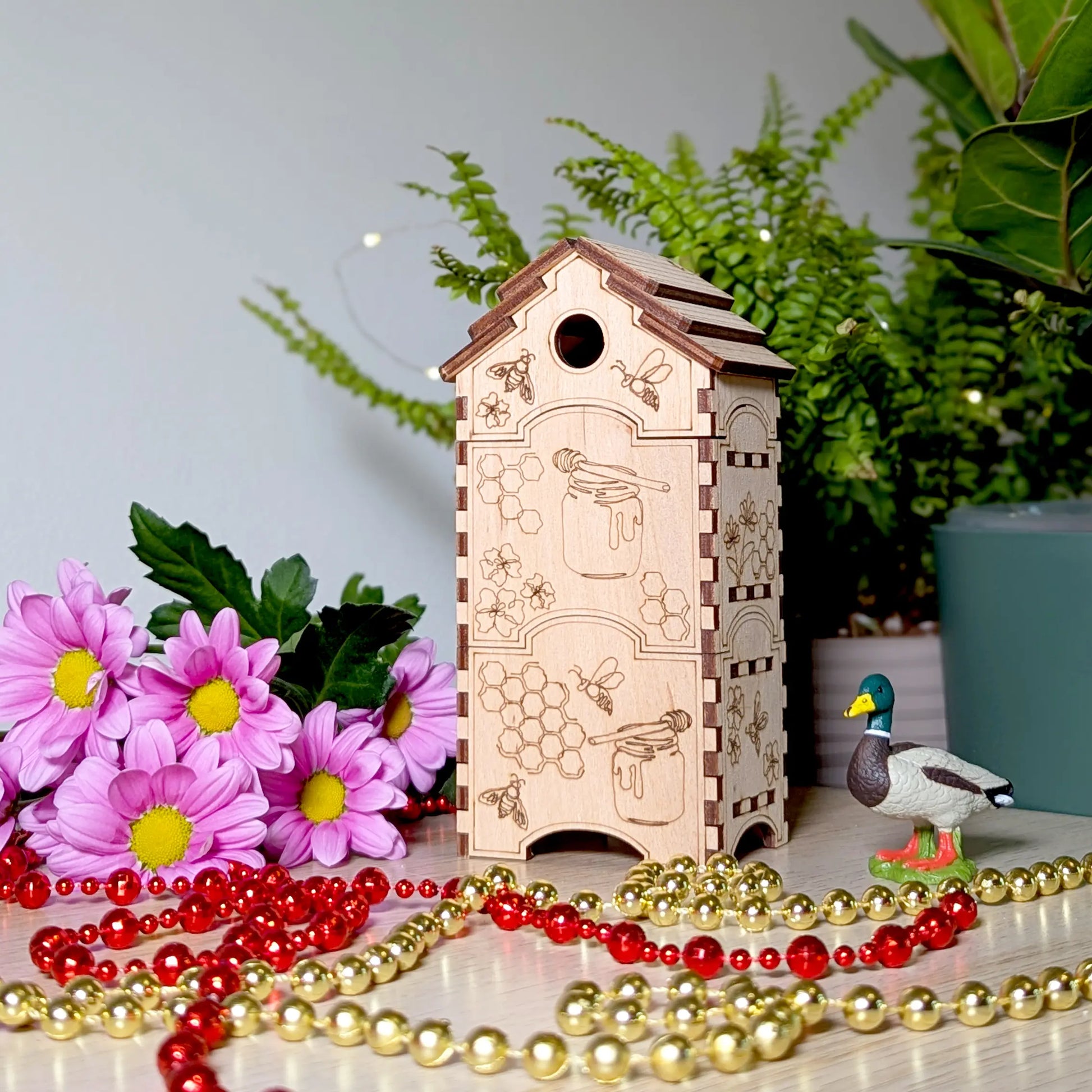 Tall double-stacked bee hive box of laser cut plywood stands near pink daisies, shiny beads, and a mallard duck toy.