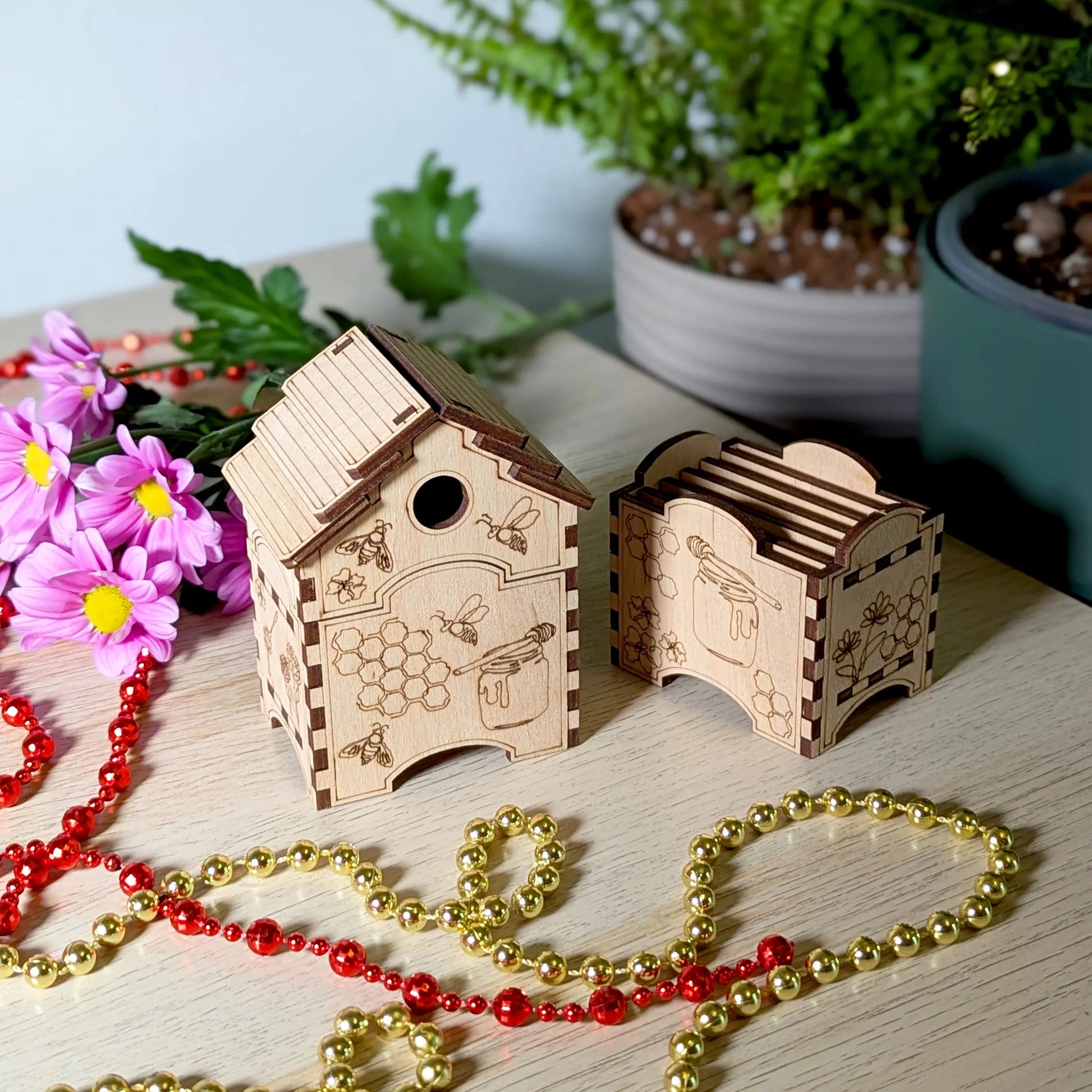 Laser cut plywood beehive box and coaster holder set staged with gold beads, pink flowers, and a potted plant.