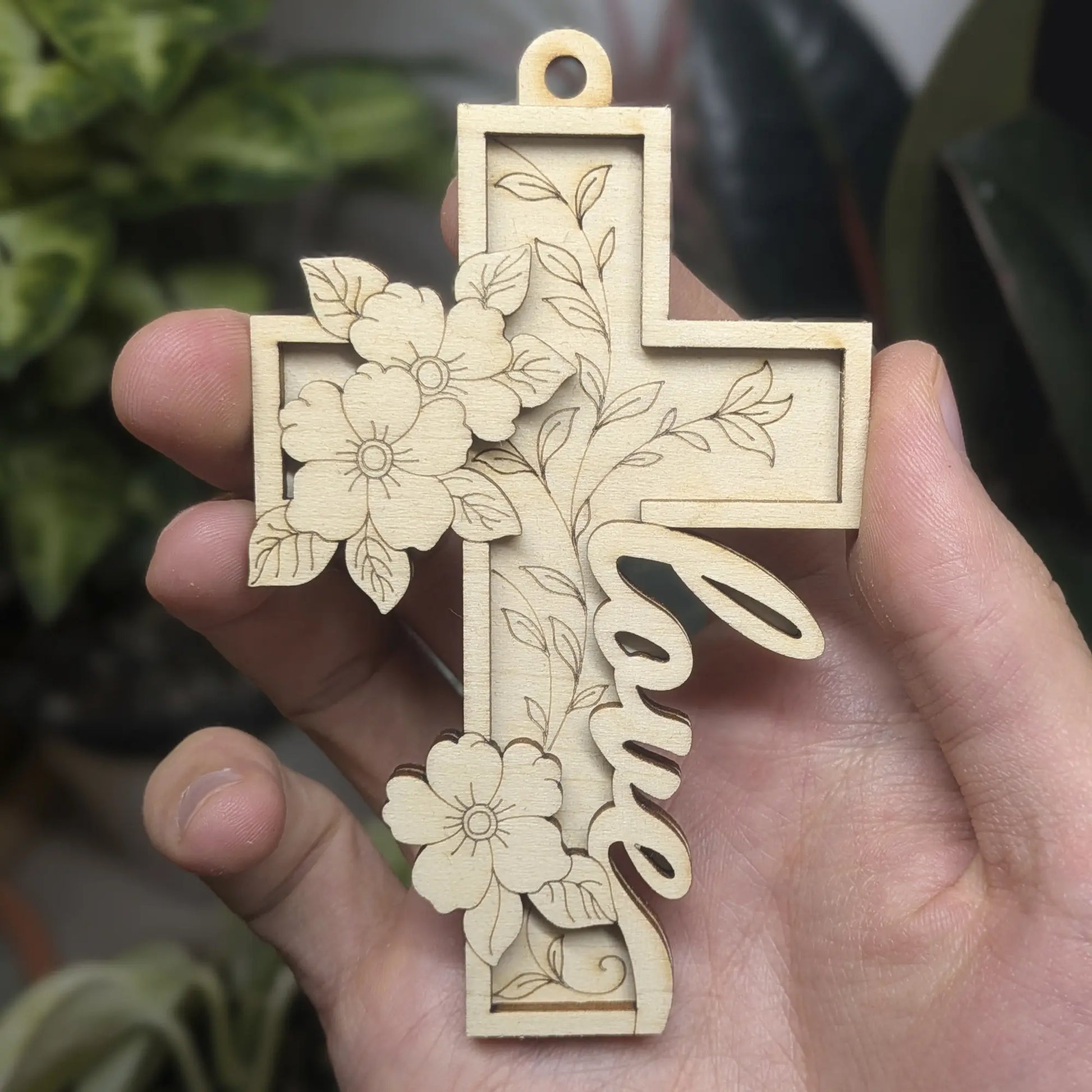 Hand holds a laser cut plywood cross ornament with the word Love in script and layered floral details. Scale is shown in hand