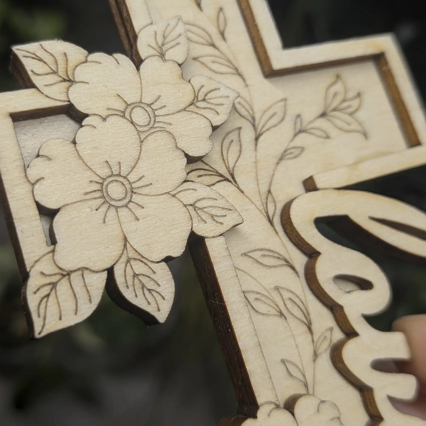 Close-up of Love script on laser cut plywood cross, showing dark burnt edges and layered floral texture in light natural wood