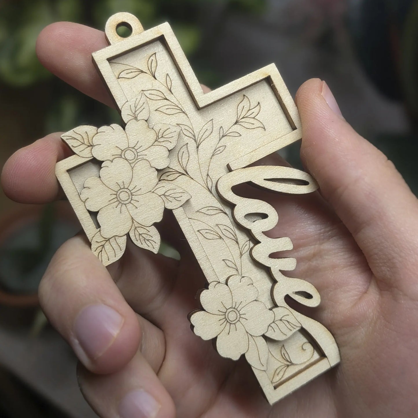 Laser cut plywood cross ornament held in hand, featuring a script Love message and intricate 3D floral branch decorations