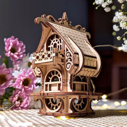 Miniature laser cut plywood house with balcony, detailed arches, and shingled roof, surrounded by floral decor.