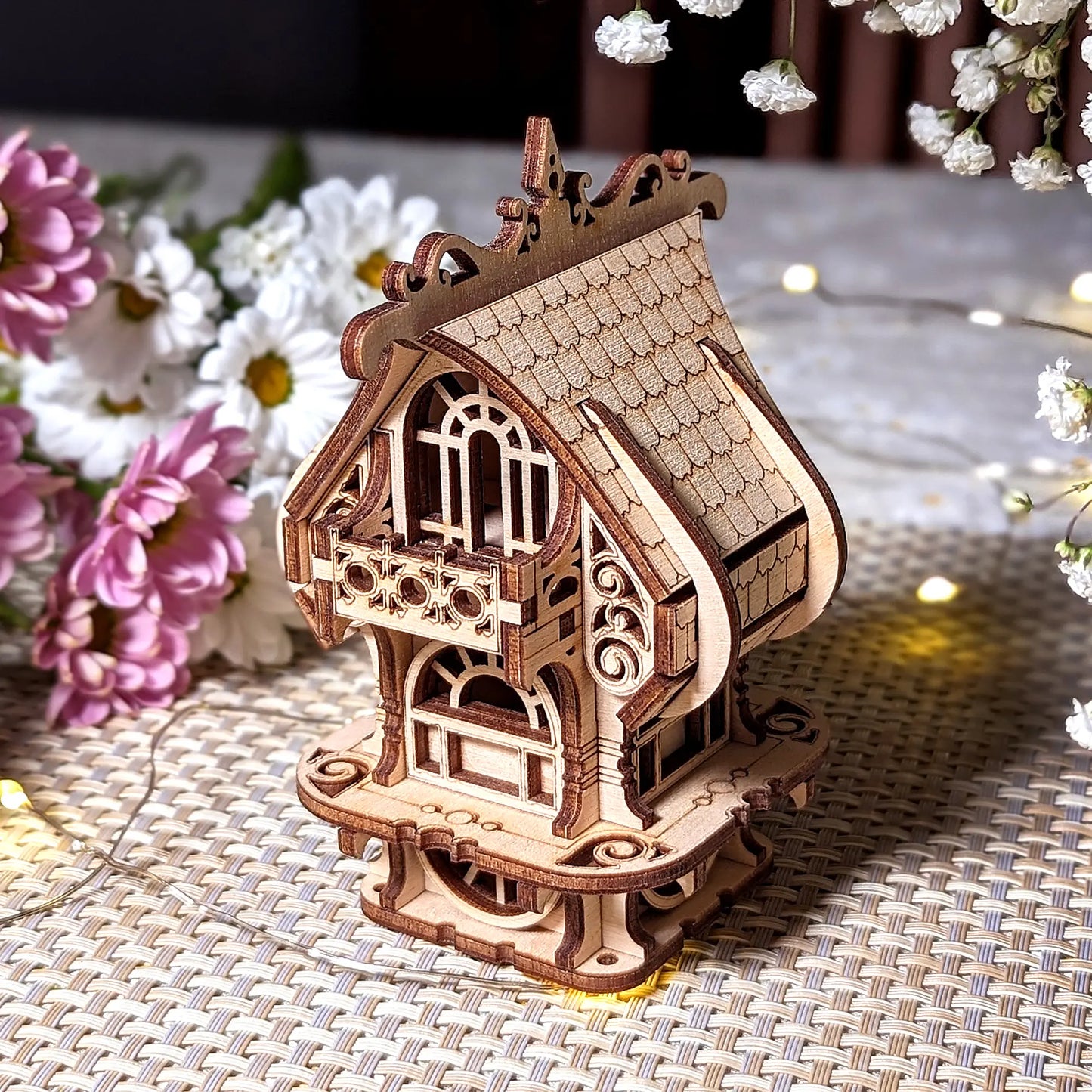 Detailed perspective of laser cut plywood house model with shingled roof and decorative balcony. Floral setting.