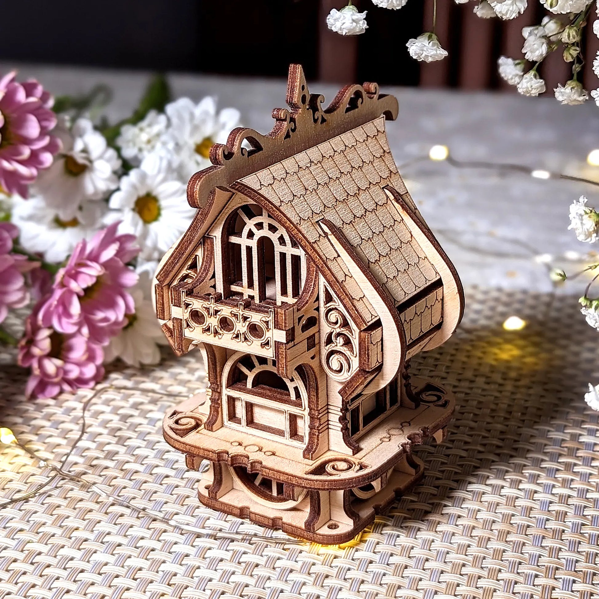 Detailed perspective of laser cut plywood house model with shingled roof and decorative balcony. Floral setting.