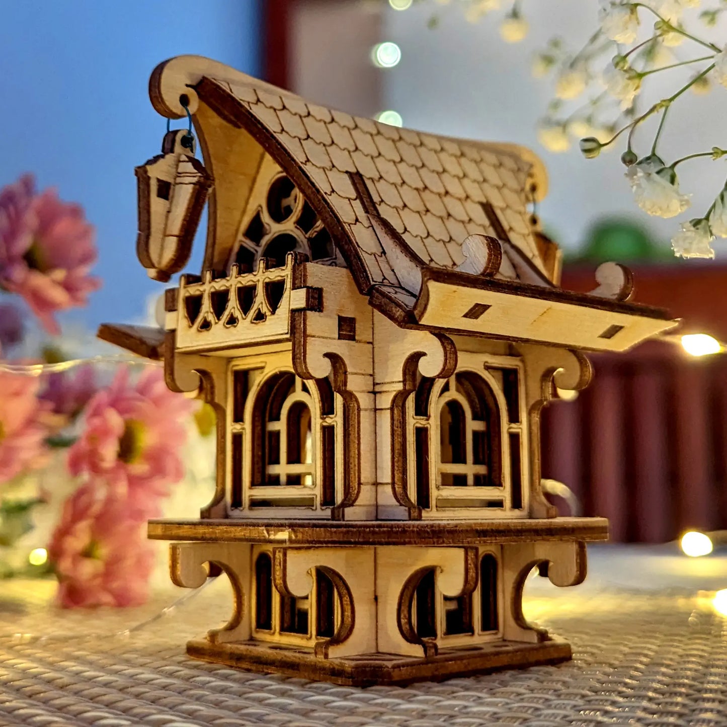 Detailed laser cut plywood fairy house on a textured mat, surrounded by soft-focus flowers and warm ambient light.