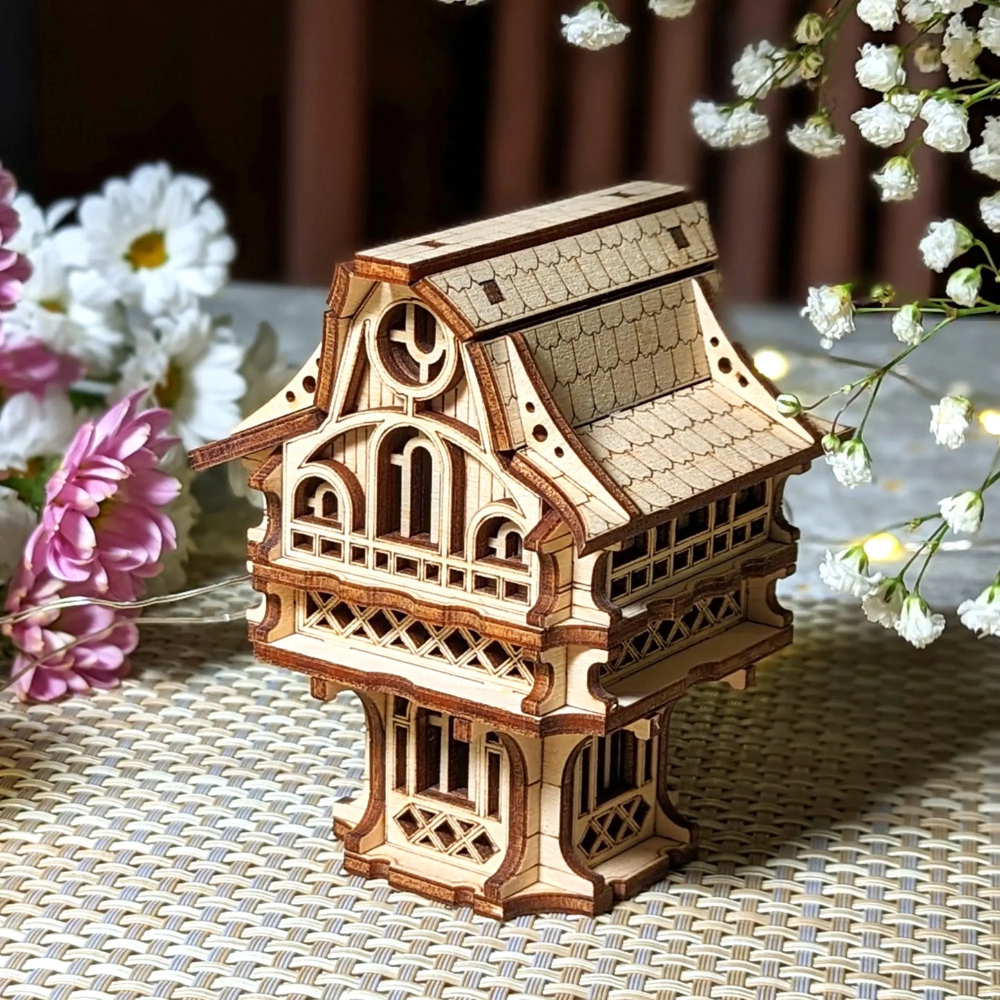 Miniature laser cut plywood cottage standing on a woven mat, beautifully staged with white gypsophila flowers and fairy lights.