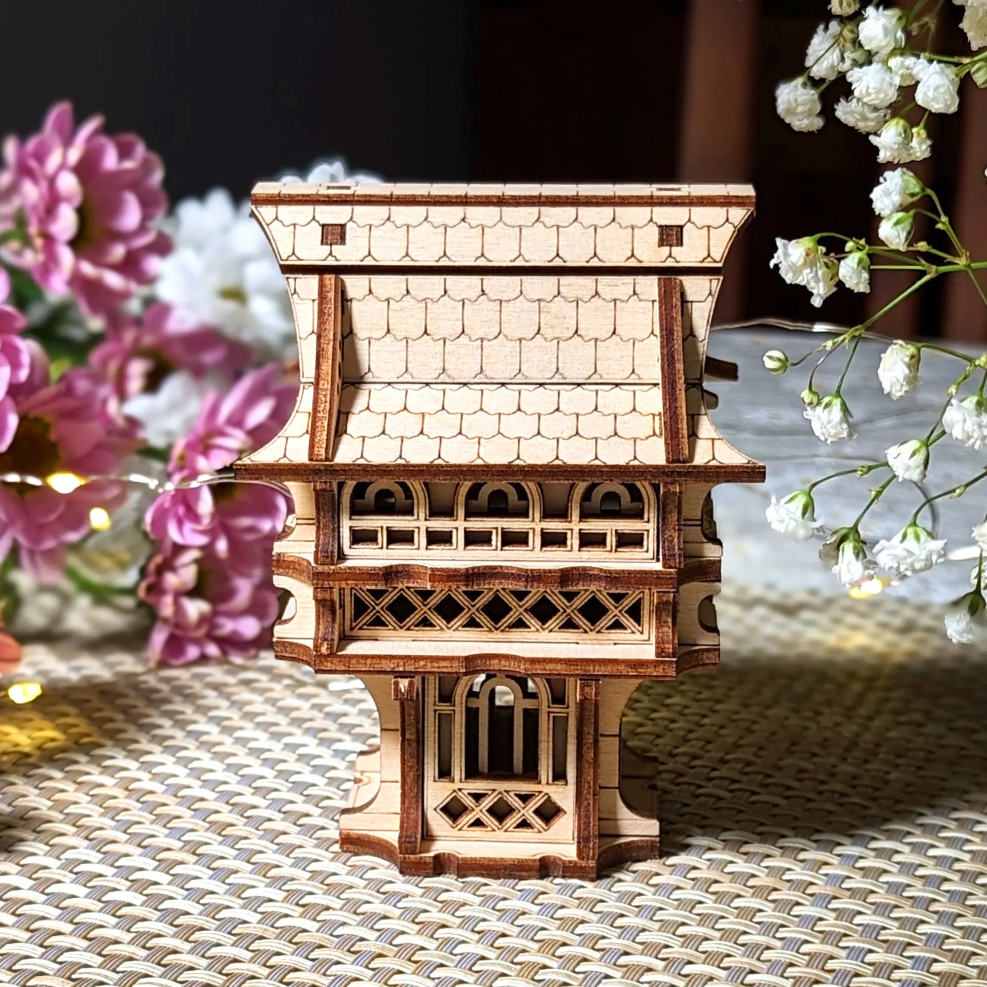 Side view of a laser cut plywood gnome house featuring a tiled roof design and ornate balcony, set against soft flowers.