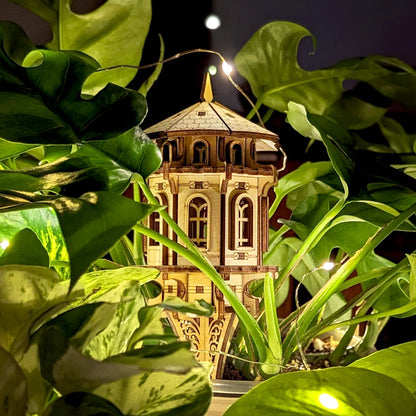 Charming laser cut plywood tower placed among indoor foliage, illuminated by warm lights, perfect for garden decor.