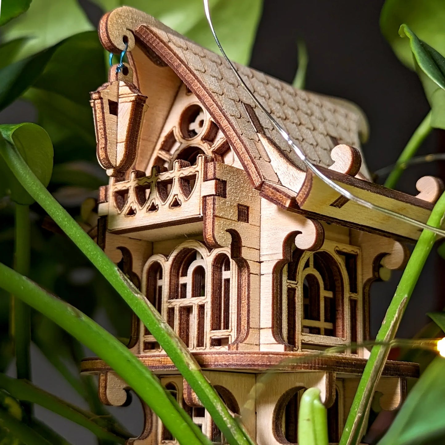 Charming laser cut plywood fairy house with hanging lantern detail, displayed amidst lush indoor foliage.