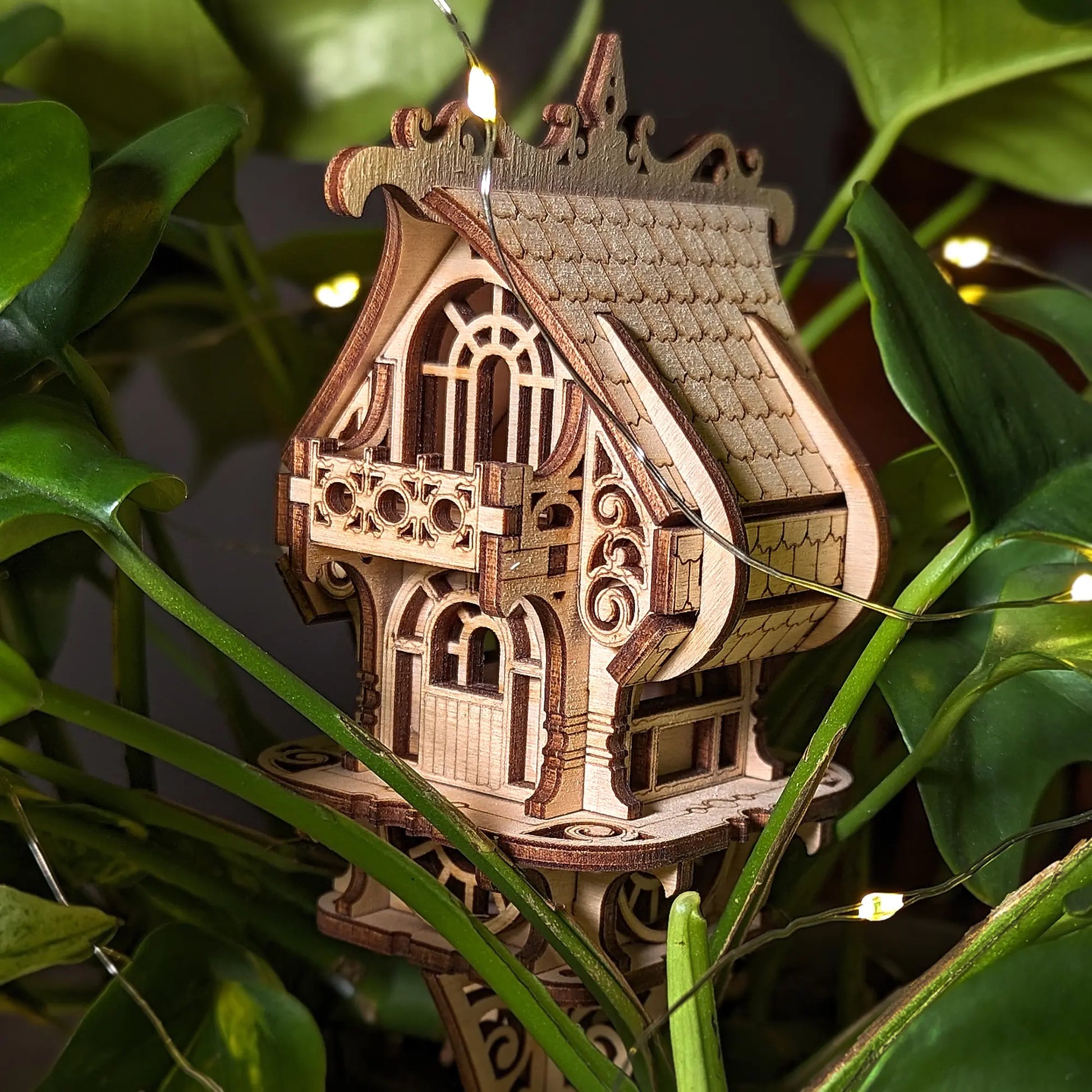 Close-up of laser cut plywood house model placed among lush green foliage and warm fairy lights. Magical style.