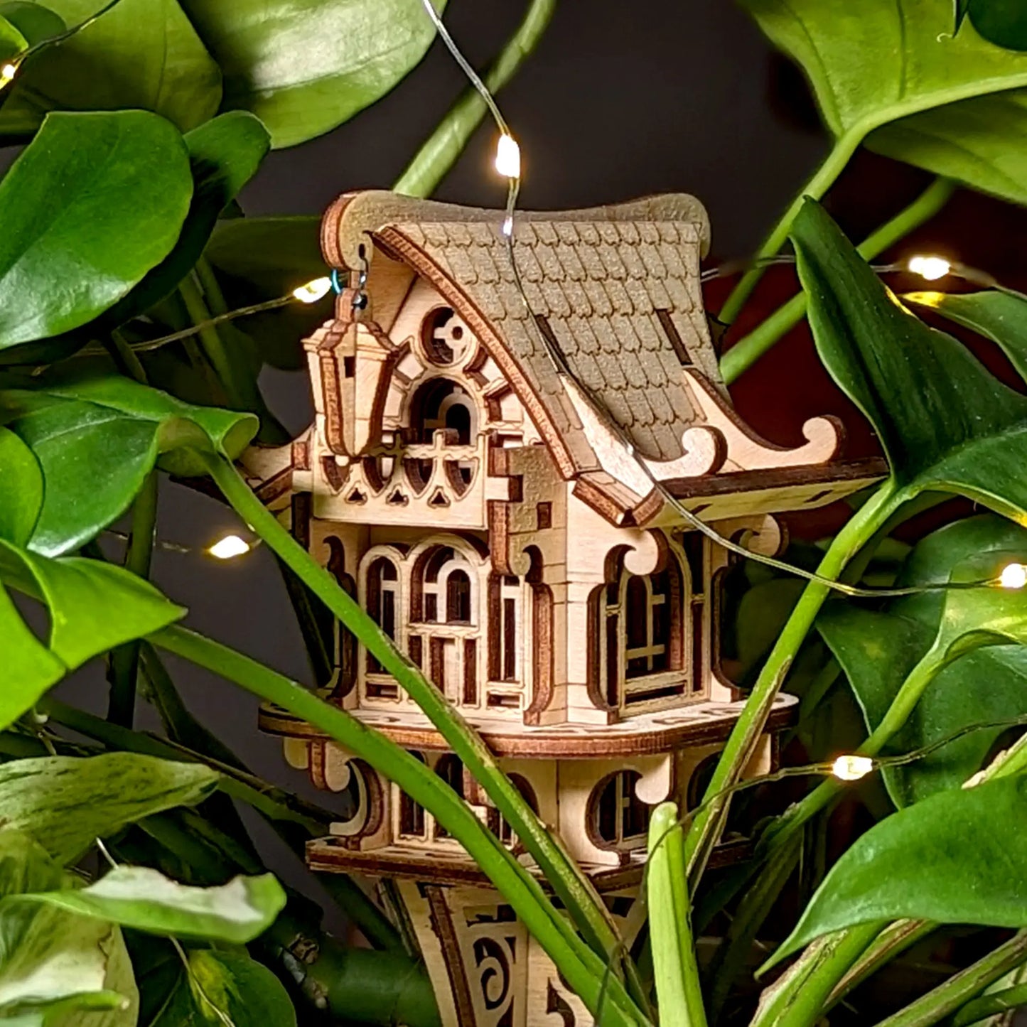 Laser cut plywood fairy house nestled in green leaves, accented by warm fairy lights for an enchanted garden look.
