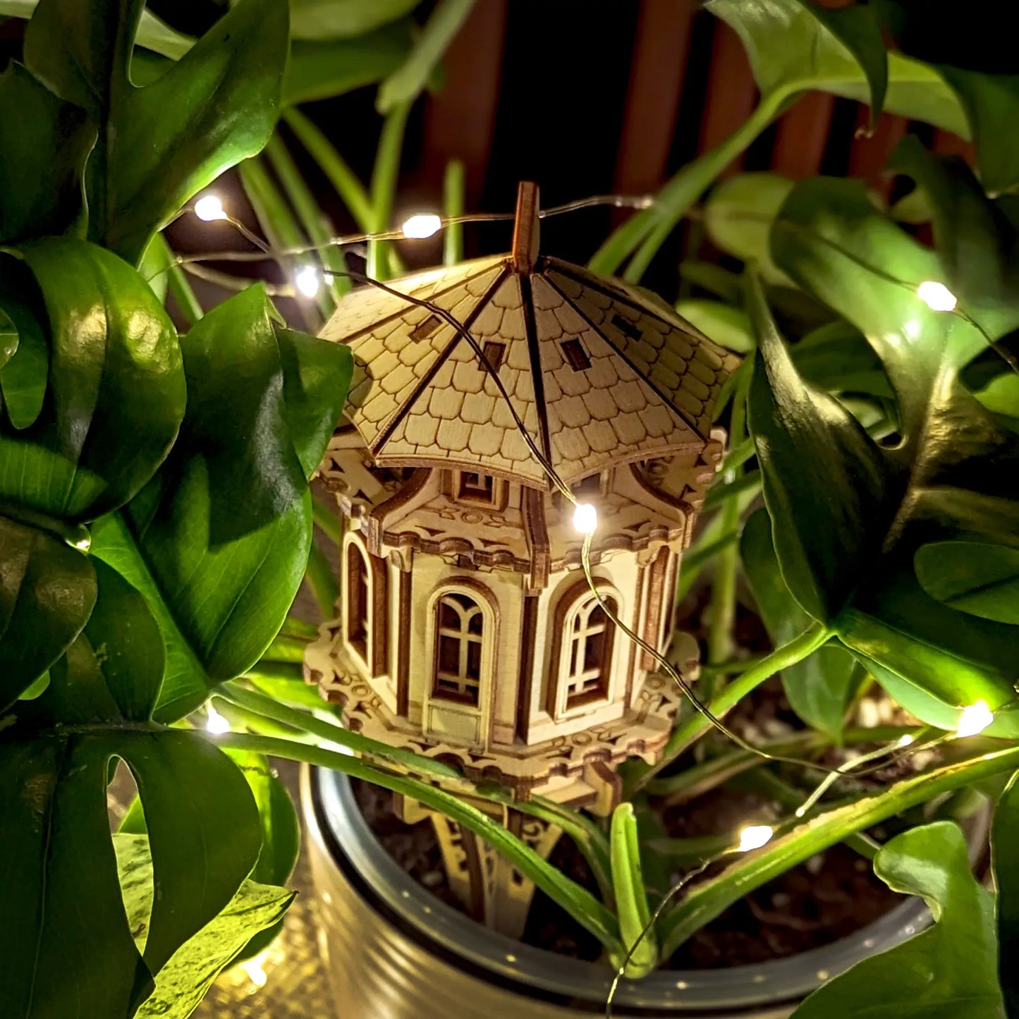 Decorative laser cut plywood garden tower staked in a plant pot, surrounded by lush leaves and glowing fairy lights.